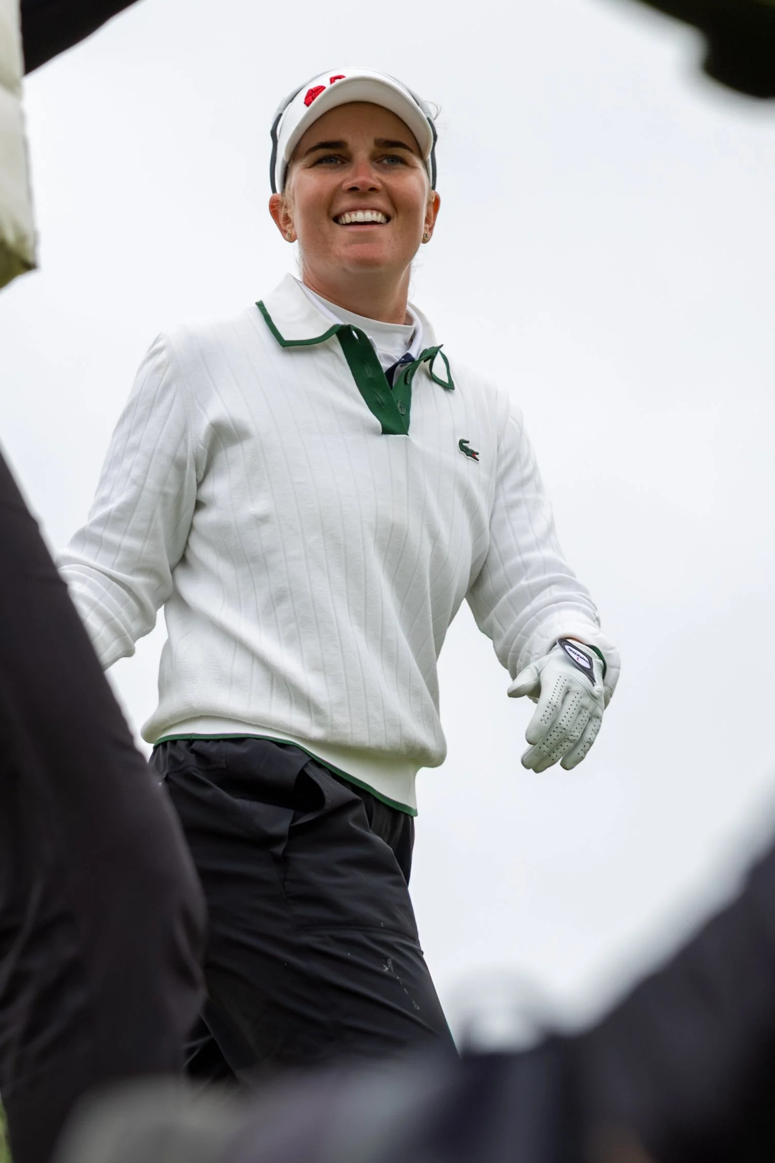 Smiling female golfer wearing a white Lacoste sweater, white cap, and golf glove, standing outdoors against a cloudy sky.