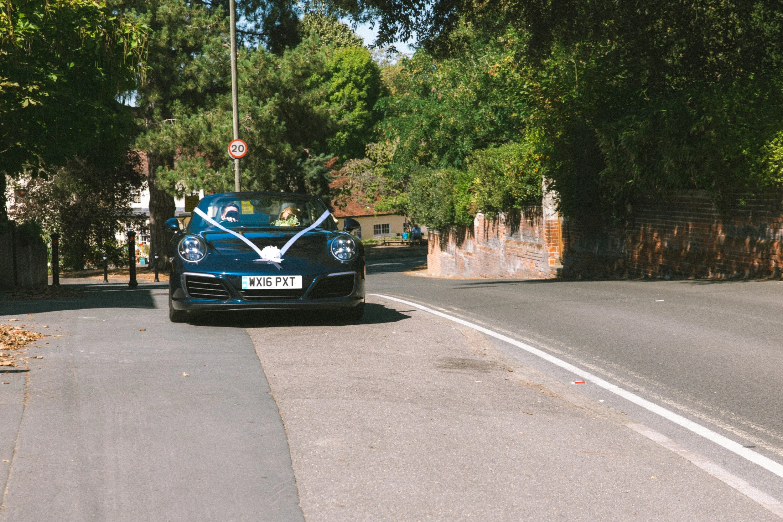 A black sports car decorated with white ribbons and a flower, driving down a leafy suburban street, possibly for a wedding.