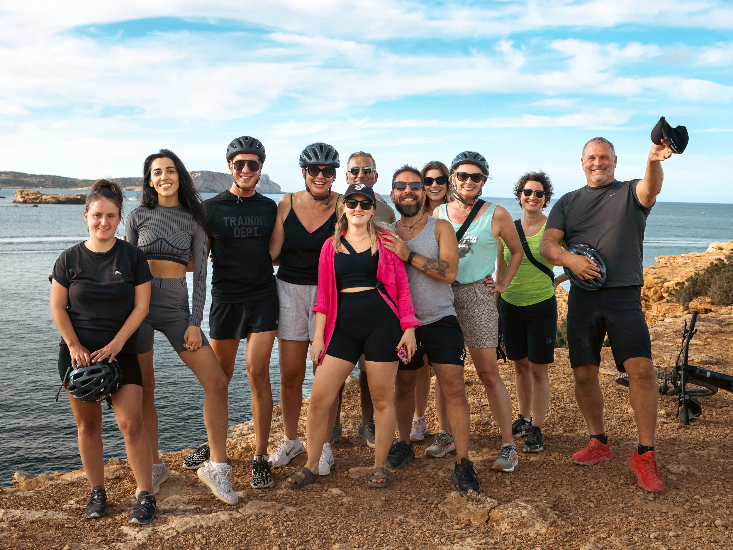 A group of twelve people, wearing casual and athletic clothing, some with bike helmets and sunglasses, stand on a rocky cliffside by the ocean, smiling and posing for a photo on a sunny day.