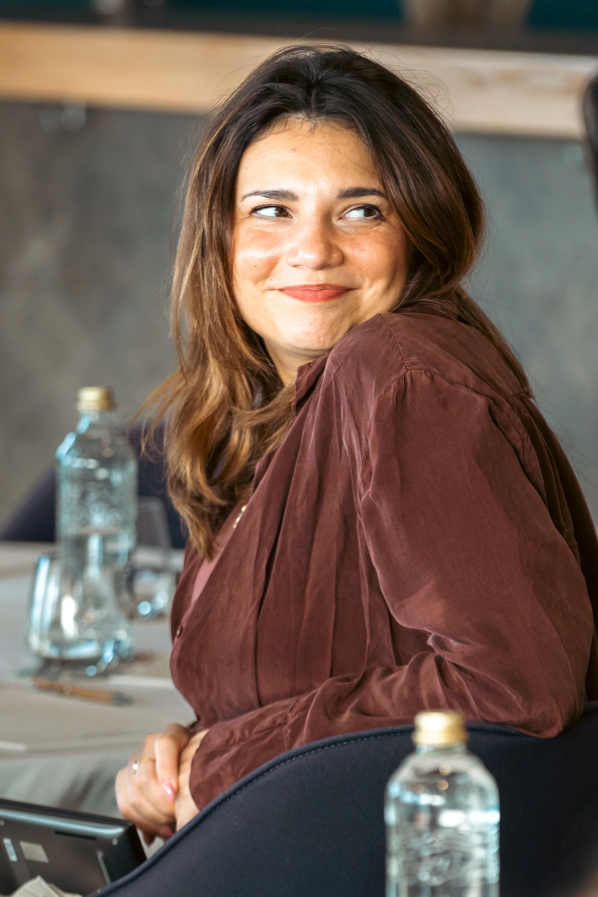 A woman with shoulder-length brown hair smiling and looking to her right, sitting at a table with a water bottle in front of her.