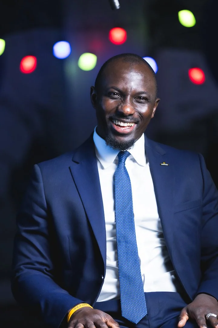 A smiling man wearing a navy blue suit, white shirt, and blue tie sitting in front of colorful blurred lights.