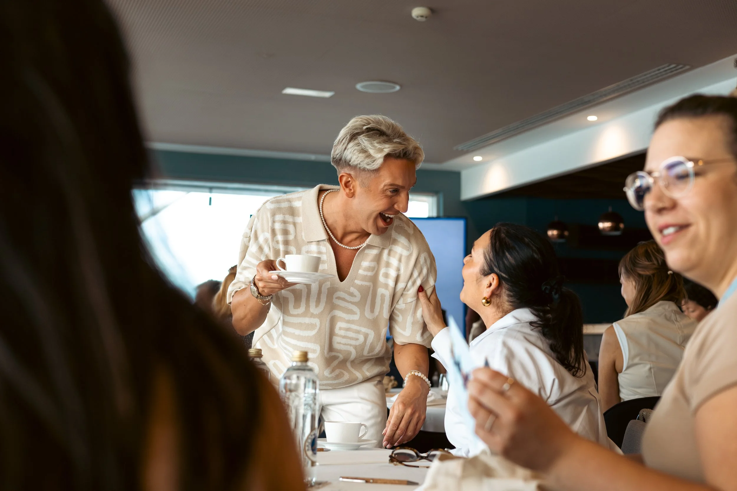 A woman with short blonde hair smiling and holding a coffee cup is talking to another woman in a white blazer in a meeting or conference room. Several other women are seated at the table, with some glasses and bottles visible on the table. The room h