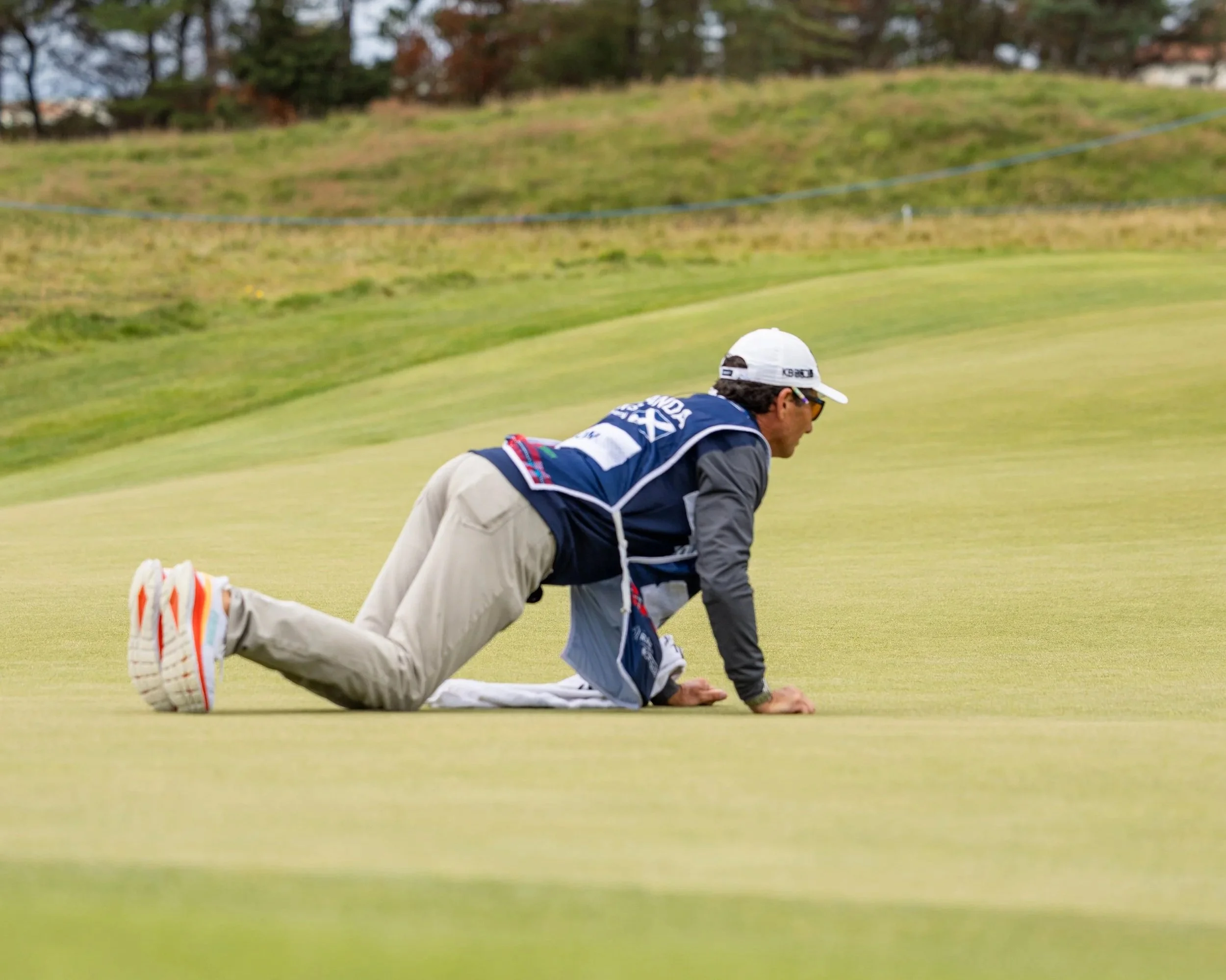 A person crawling on the green grass of a golf course, wearing a navy jacket, beige pants, white sneakers with orange and blue accents, a white cap, and sunglasses. Background shows a grassy hill and trees.