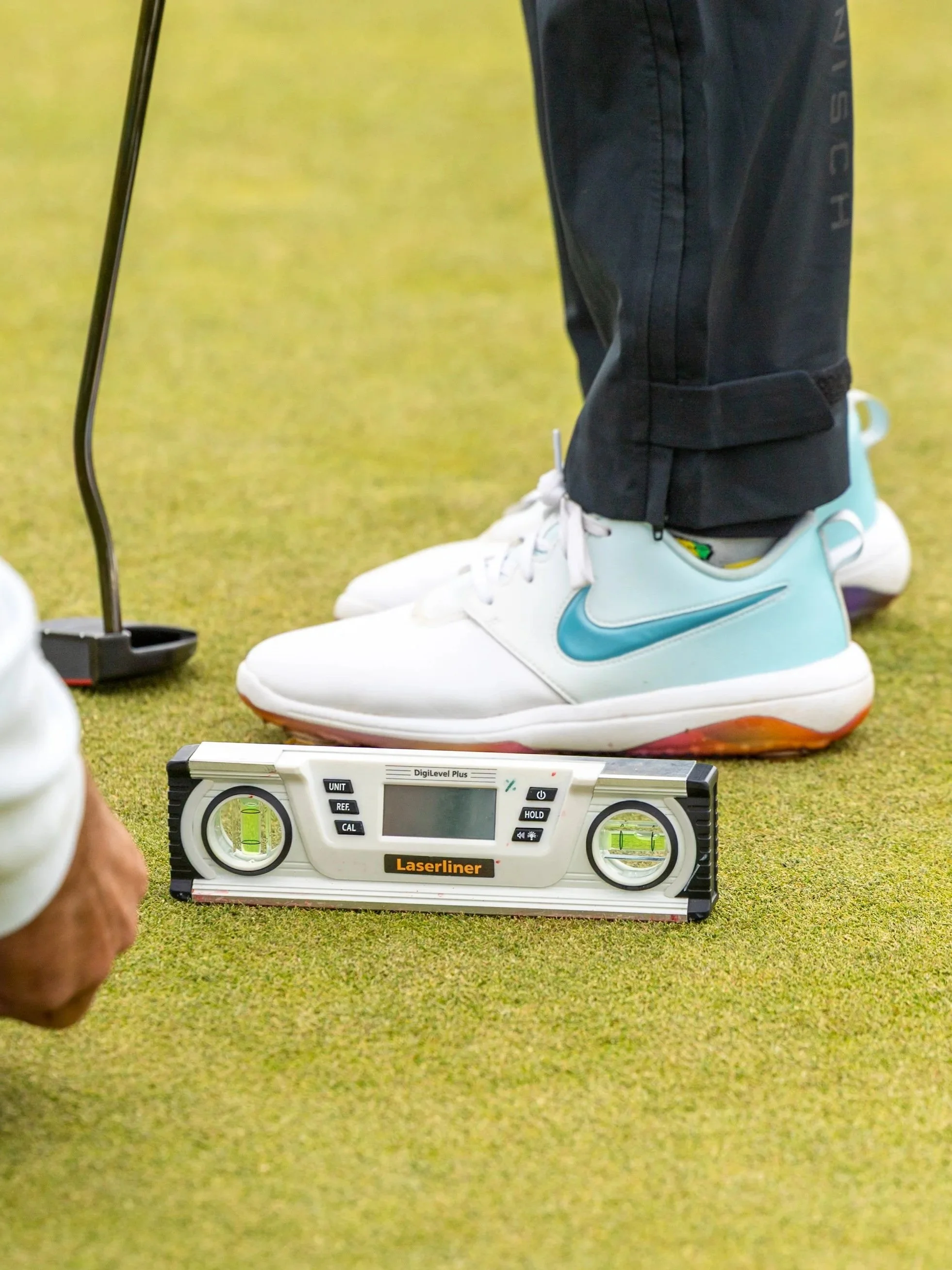 Close-up of a person wearing black pants and white golf shoes with a blue Nike swoosh, standing on a green artificial turf. In front of their feet is a laser level device with a digital display, and to the left, part of a golf club is visible.