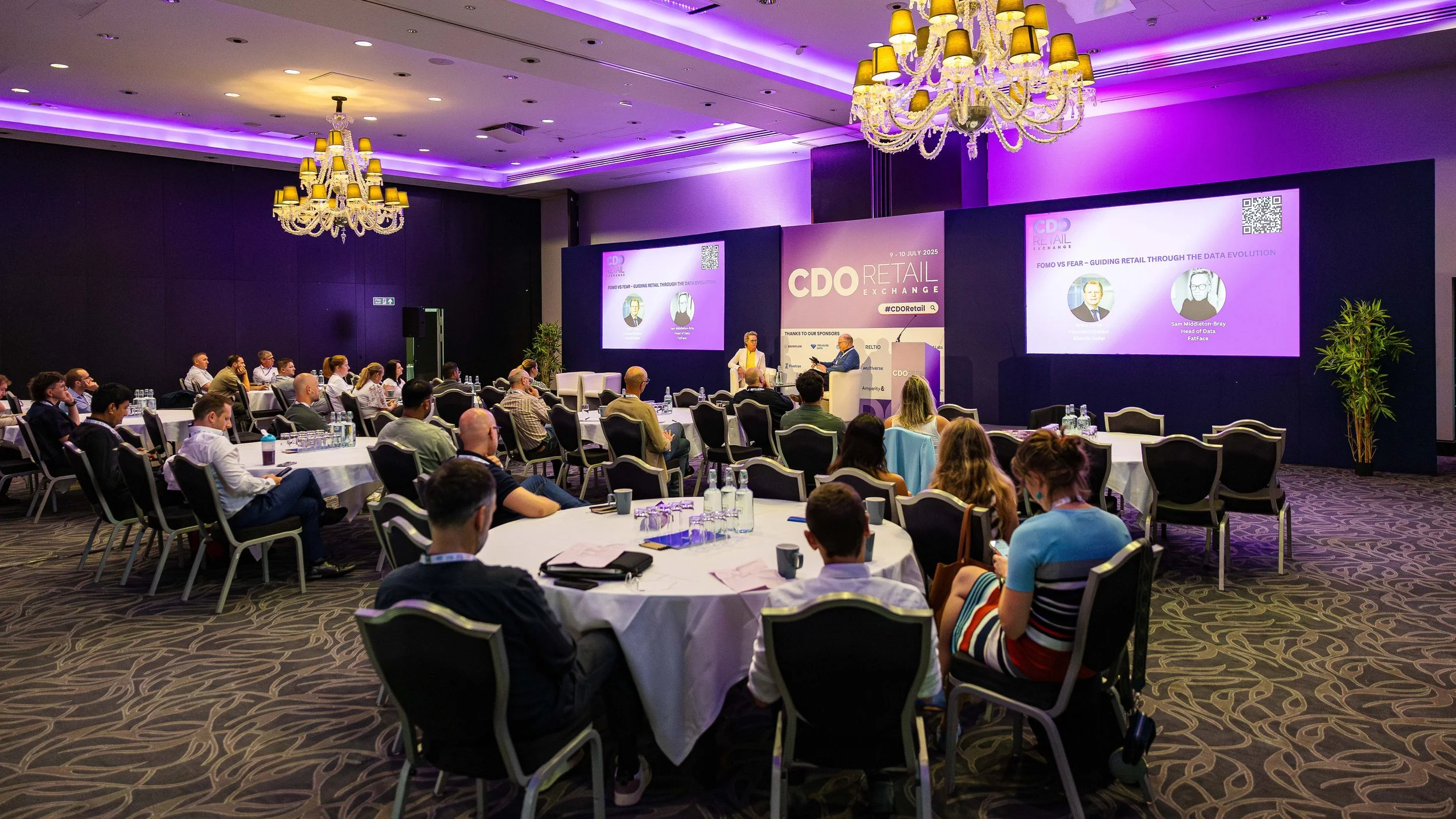 A large conference room filled with attendees seated at round tables, facing a stage with two large screens displaying a presentation on retail data. The room has elegant chandeliers, purple lighting, and dark walls.