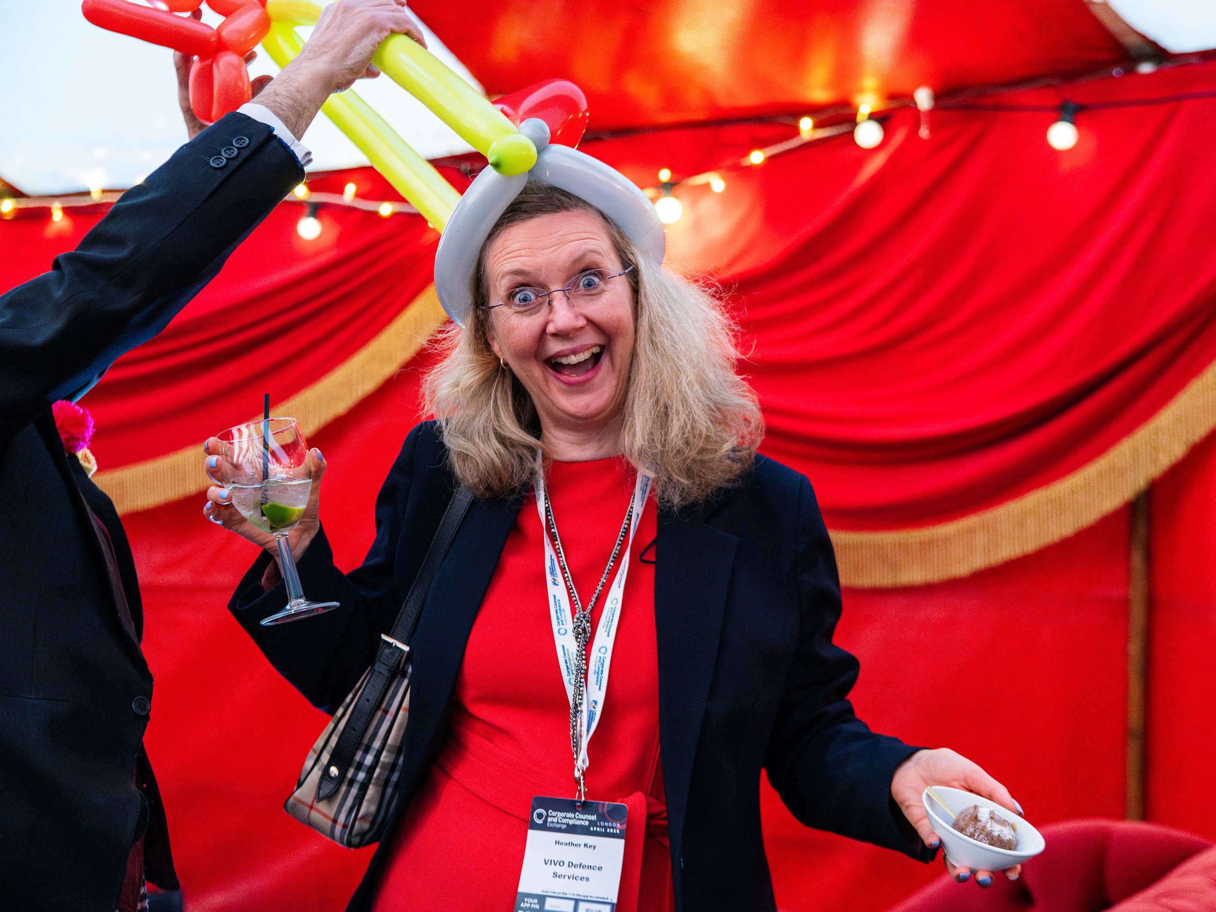 A woman with long blonde hair, glasses, and a red dress, wearing a white hat made of balloons, is smiling and holding a glass of a cocktail with lime. She is at an event with a red and yellow festive backdrop. A man in a dark suit is holding a bowl o
