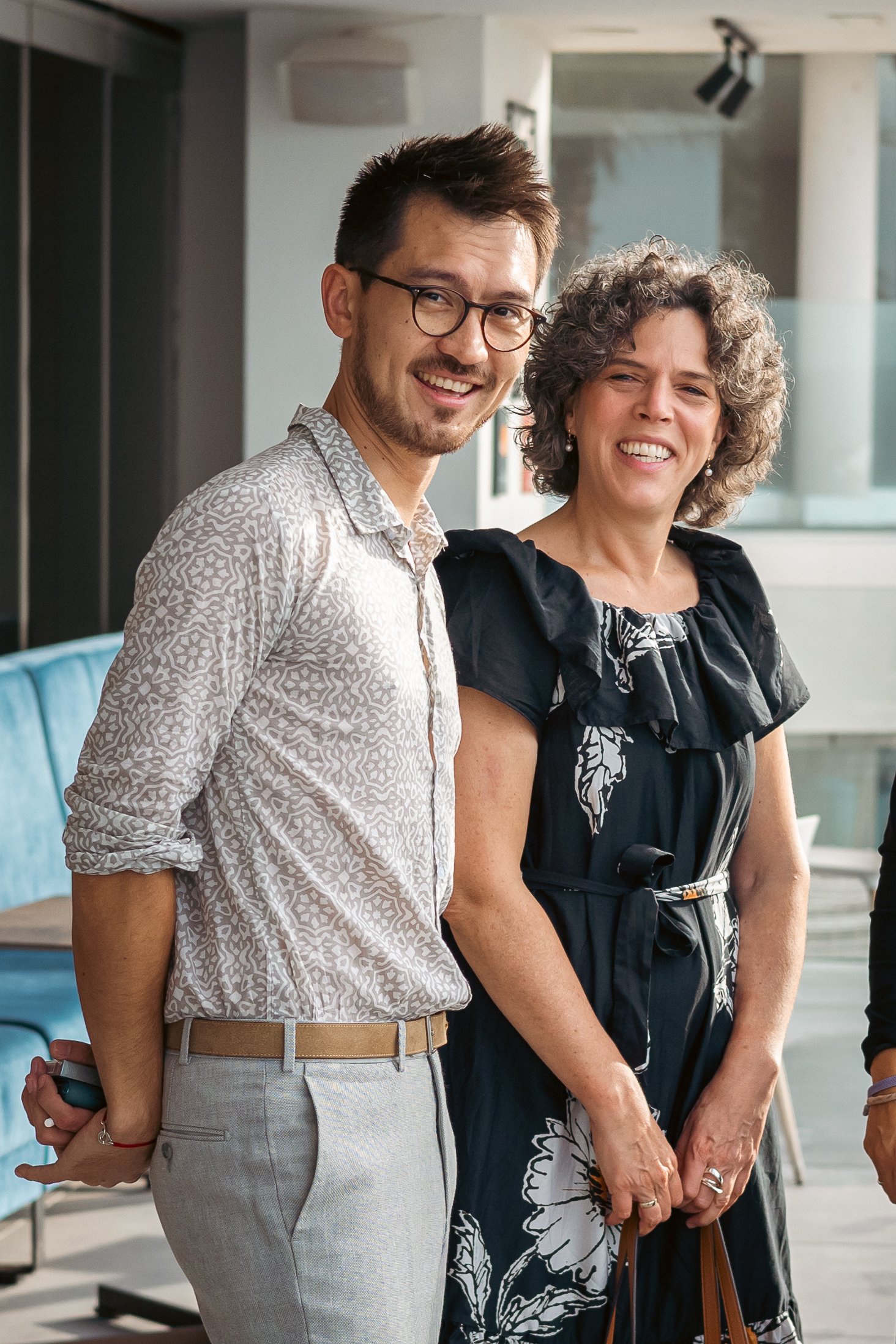 A man with glasses and a woman with curly gray hair stand close together, smiling indoors with modern decor and a large window in the background.