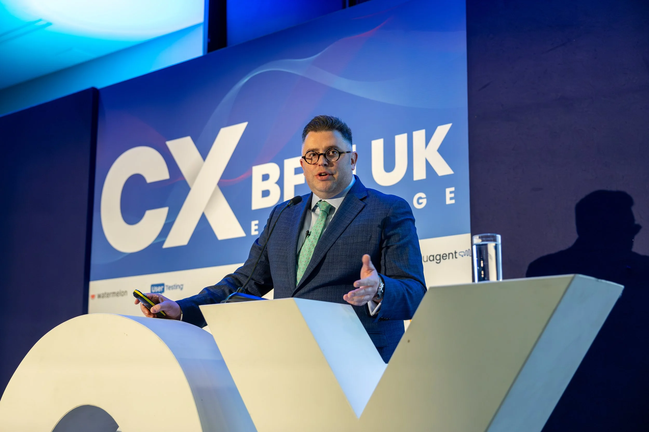 A man in a suit and glasses speaking at a conference with a large blue screen behind him displaying the words 'CX UK'. He is gesturing with his hands and holding a phone in his right hand. There is a glass of water on the table in front of him.