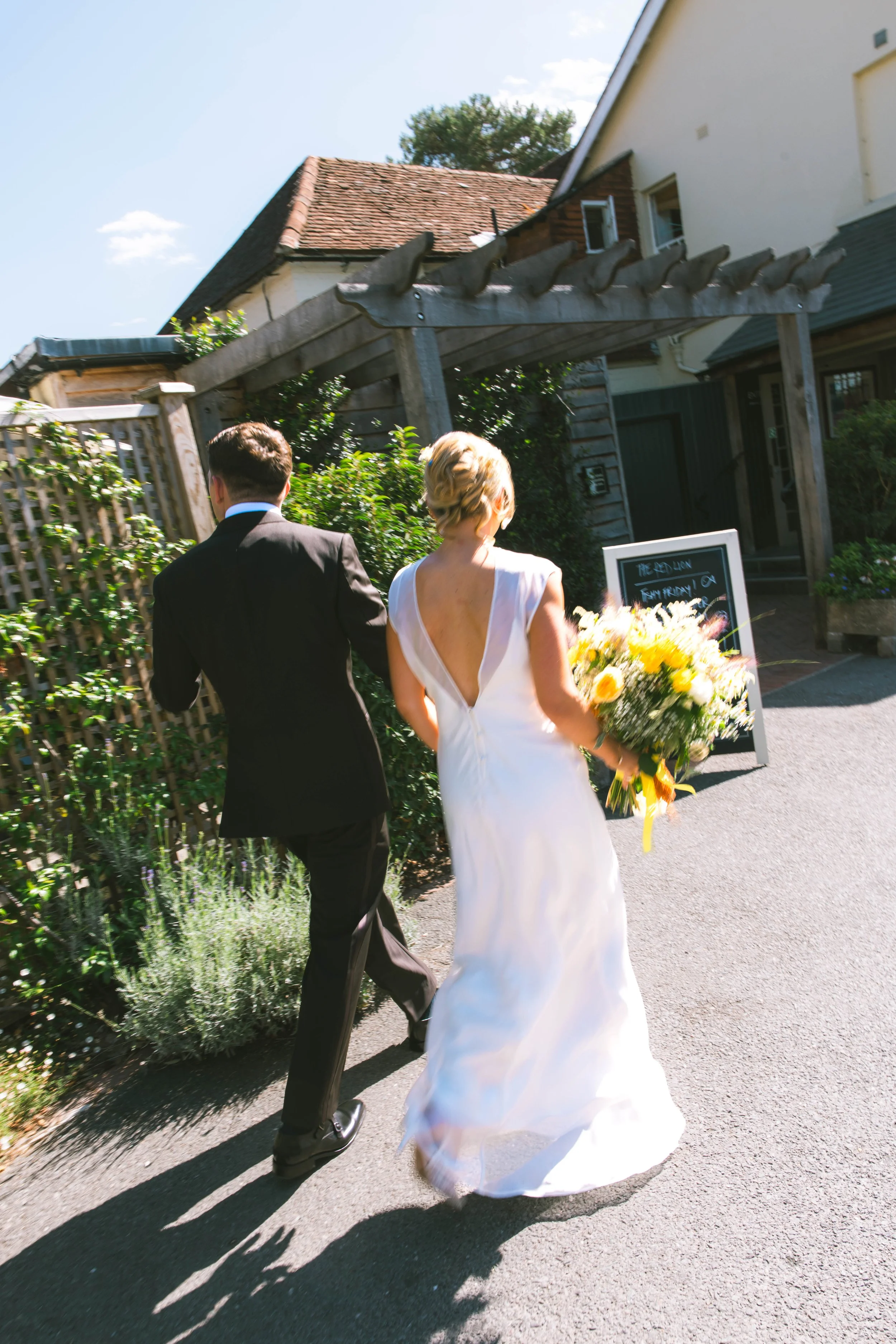 A bride in a white wedding dress holding a bouquet and a groom in a black suit walking on a sunny street outside a house with a wooden structure and a chalkboard sign nearby.