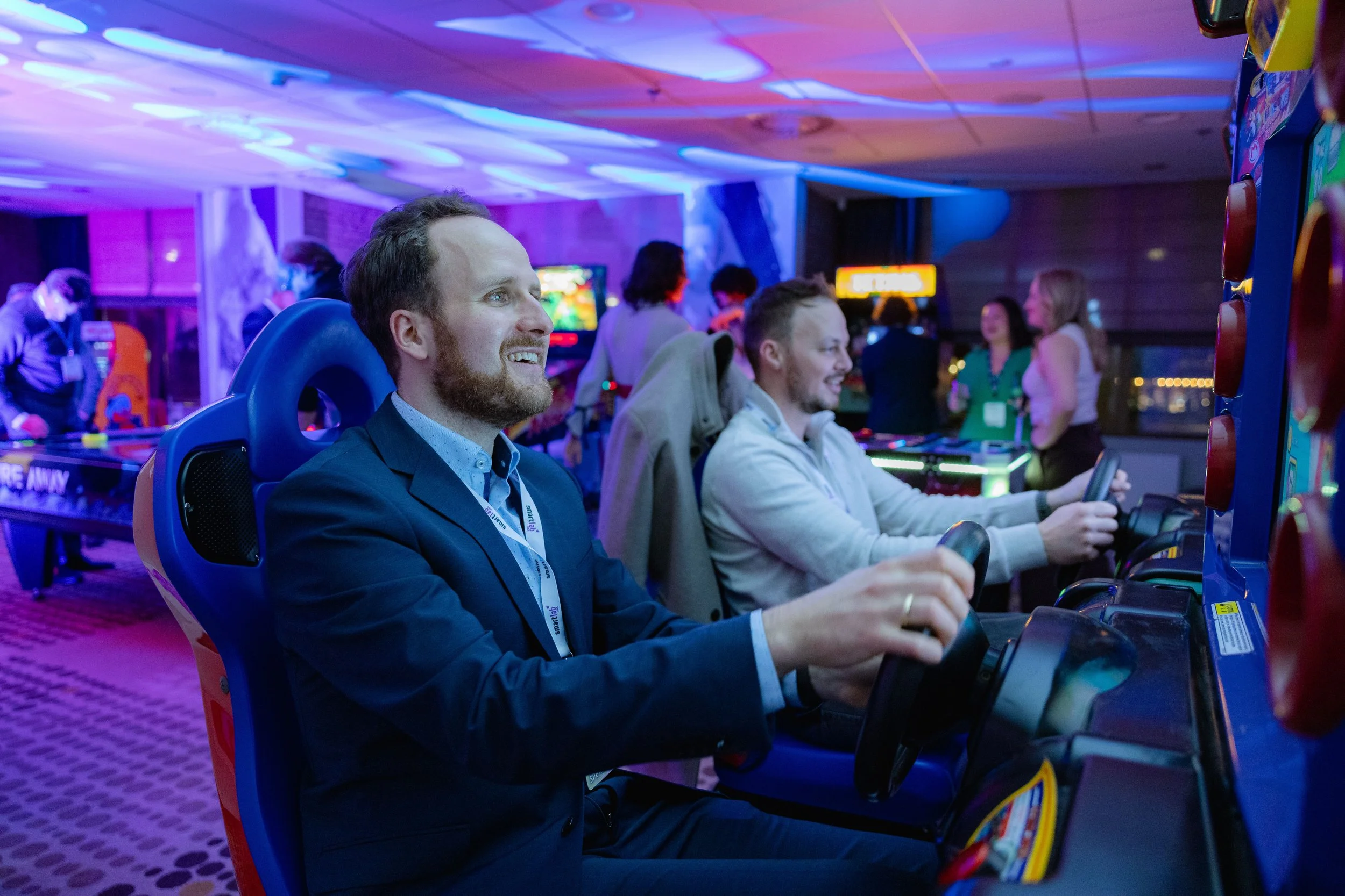 Two men playing a video arcade game in a dimly lit, colorful gaming arcade with other people in the background.