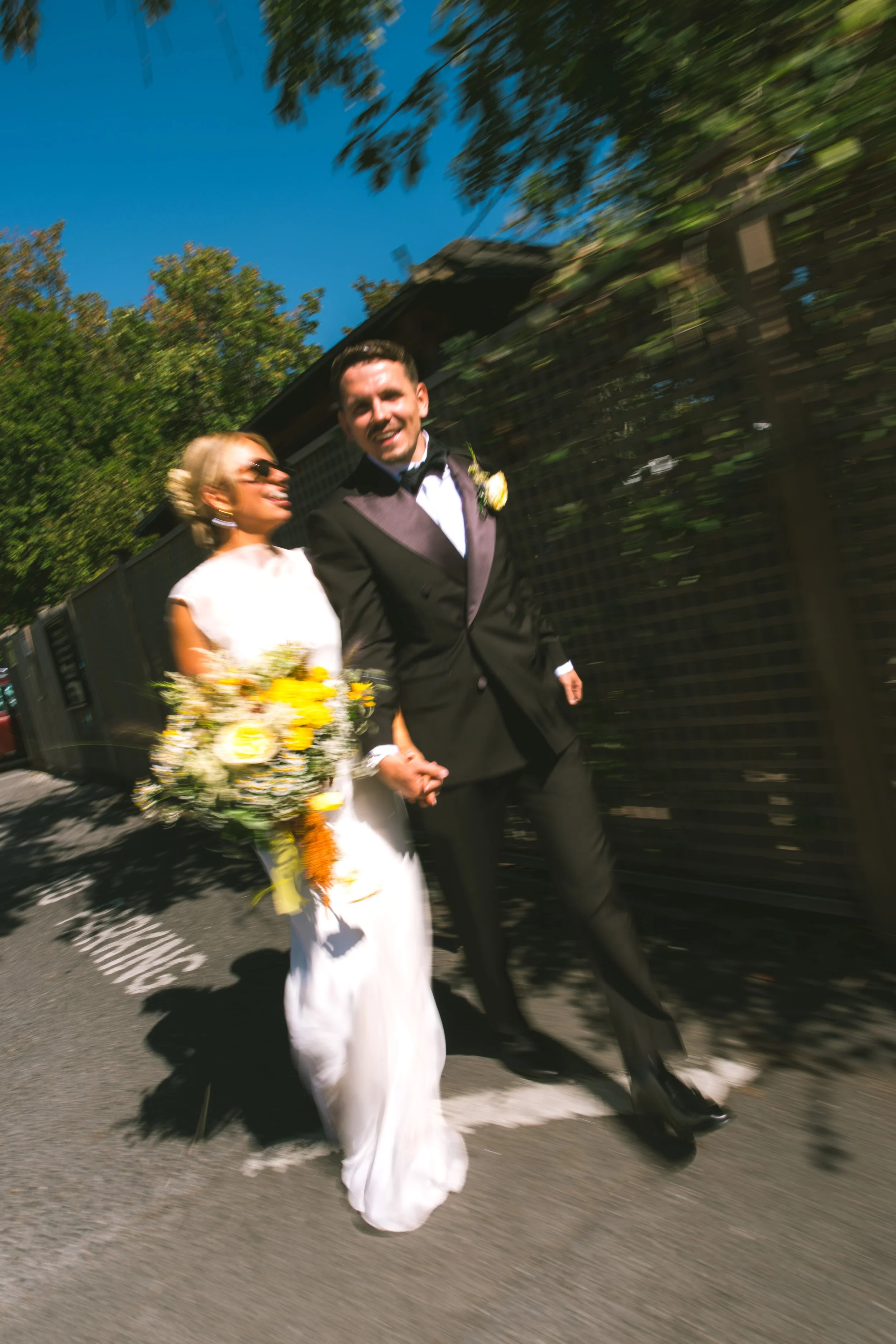 A smiling bride and groom holding hands walk down the street during a wedding celebration on a sunny day. The bride is holding a flower bouquet and is dressed in a white gown, while the groom is wearing a black tuxedo with a bow tie. There is a blurr