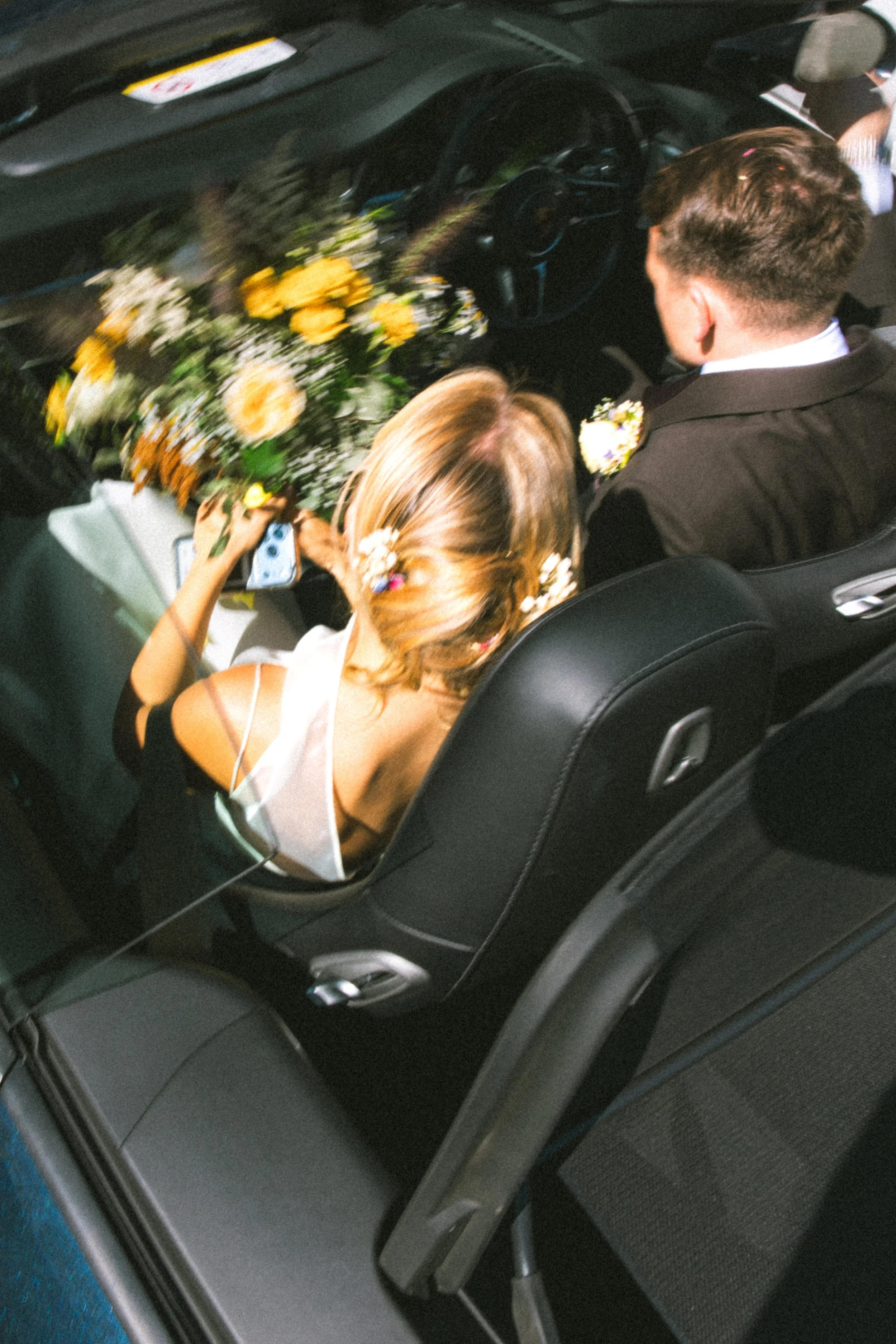 Inside a car, a man and woman are seated in the back. The woman is holding a bouquet of yellow and white flowers, and the man is wearing a suit with a boutonniere. The scene suggests a wedding or special occasion.