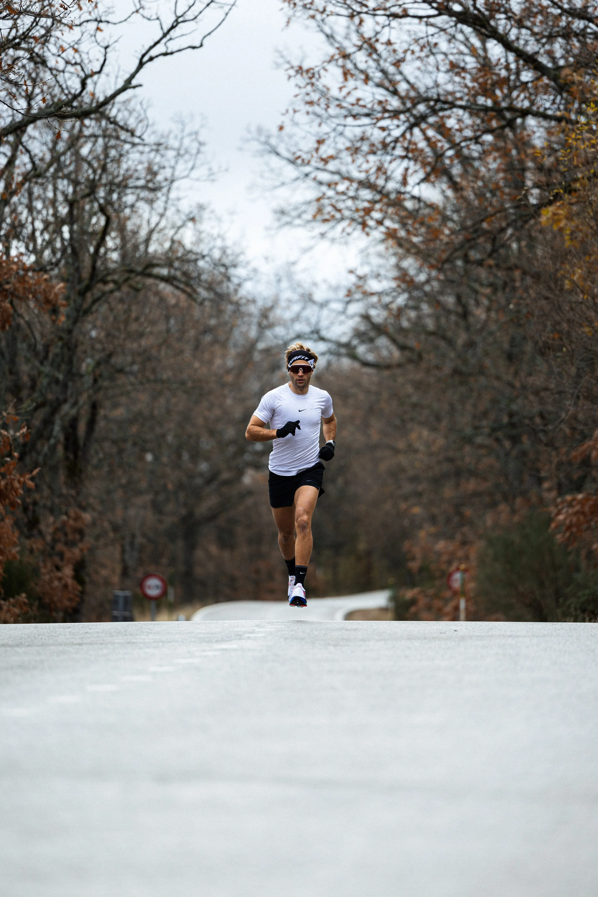 A man running on a paved road through a park with trees and autumn leaves, wearing a white shirt, black shorts, and a headband.