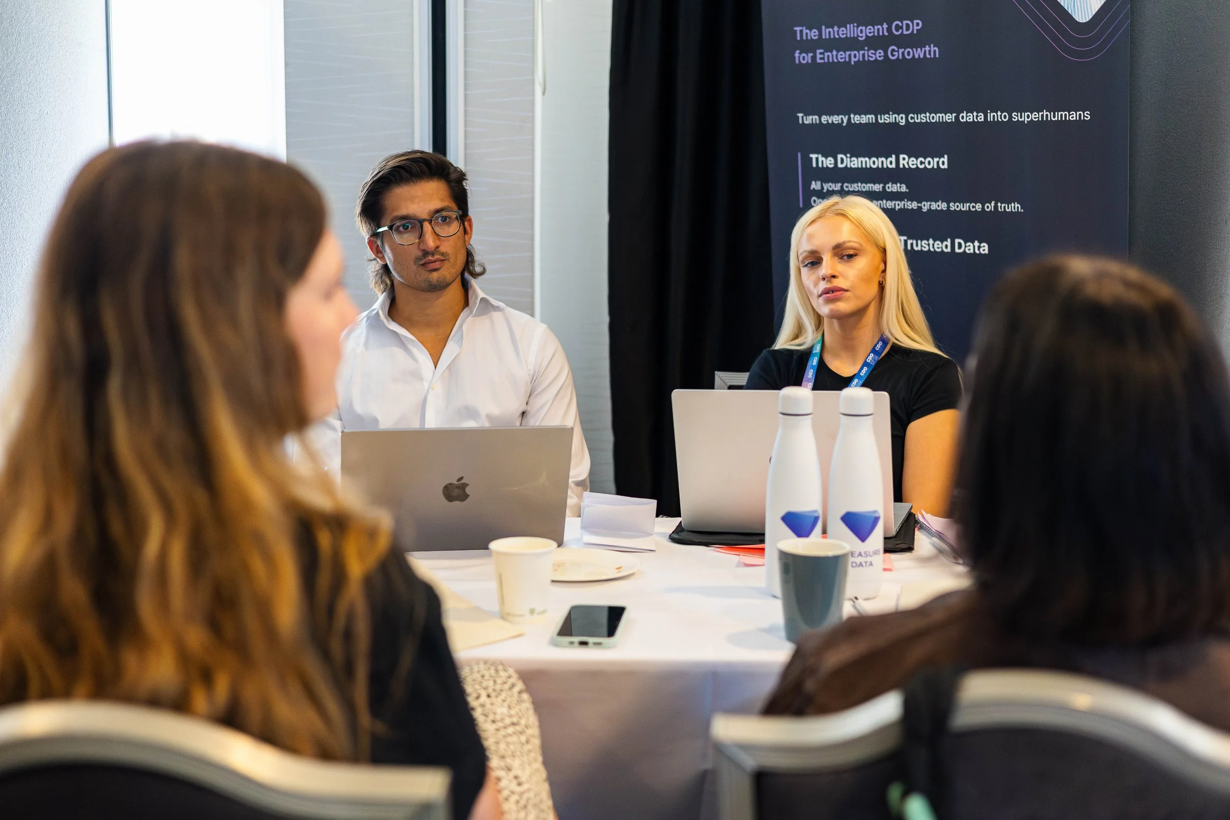 People attending a business meeting in a conference room, with a focus on a man with glasses and a woman with blonde hair, sitting at a table with laptops, water bottles, and paper cups, and a large screen displaying business information in the backg