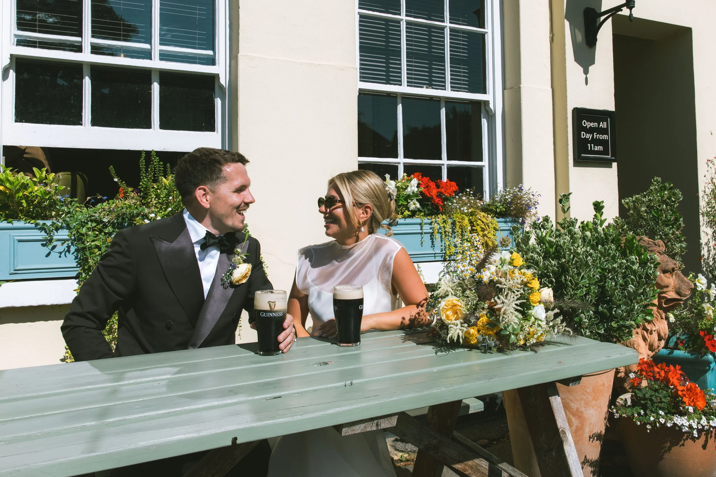 A smiling man in a black tuxedo and a woman in a white dress sitting at a table outside a building with large windows and flower boxes, holding glasses of Guinness beer, likely celebrating a special occasion.
