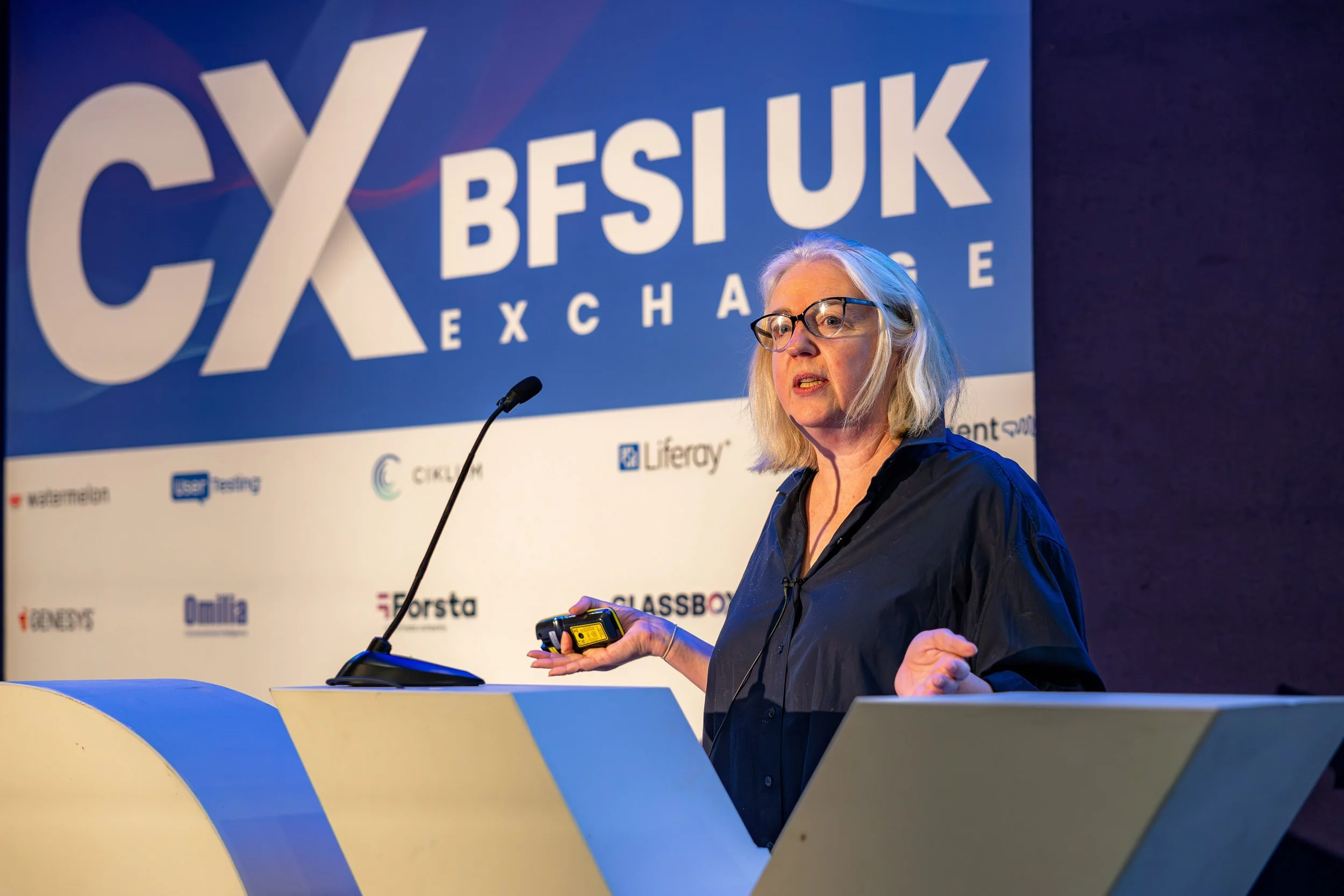 A woman wearing glasses and a dark shirt speaking at a conference. She is holding a device in her hand and standing in front of a blue backdrop with the word 'CES' and 'BESUK Exchange' along with various sponsor logos.