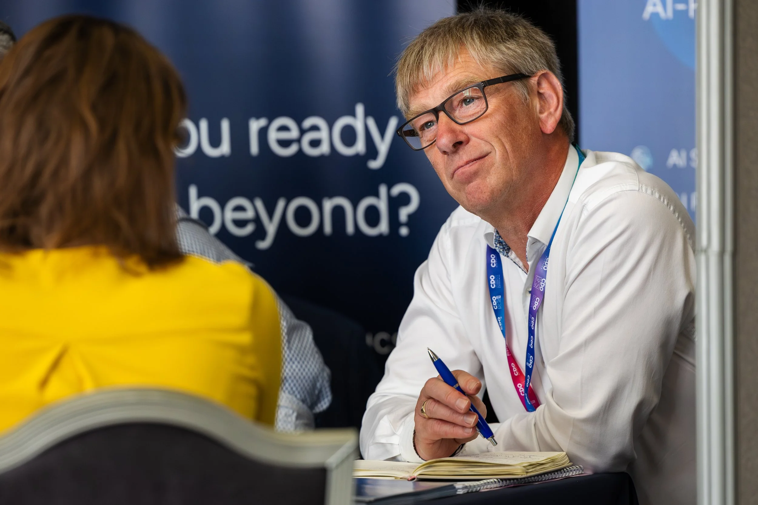 A man with glasses, light brown hair, and a white shirt is sitting at a table, looking at a woman in a yellow outfit with her back to the camera. The man has a lanyard with a badge around his neck and is holding a blue pen over a notebook. In the bac