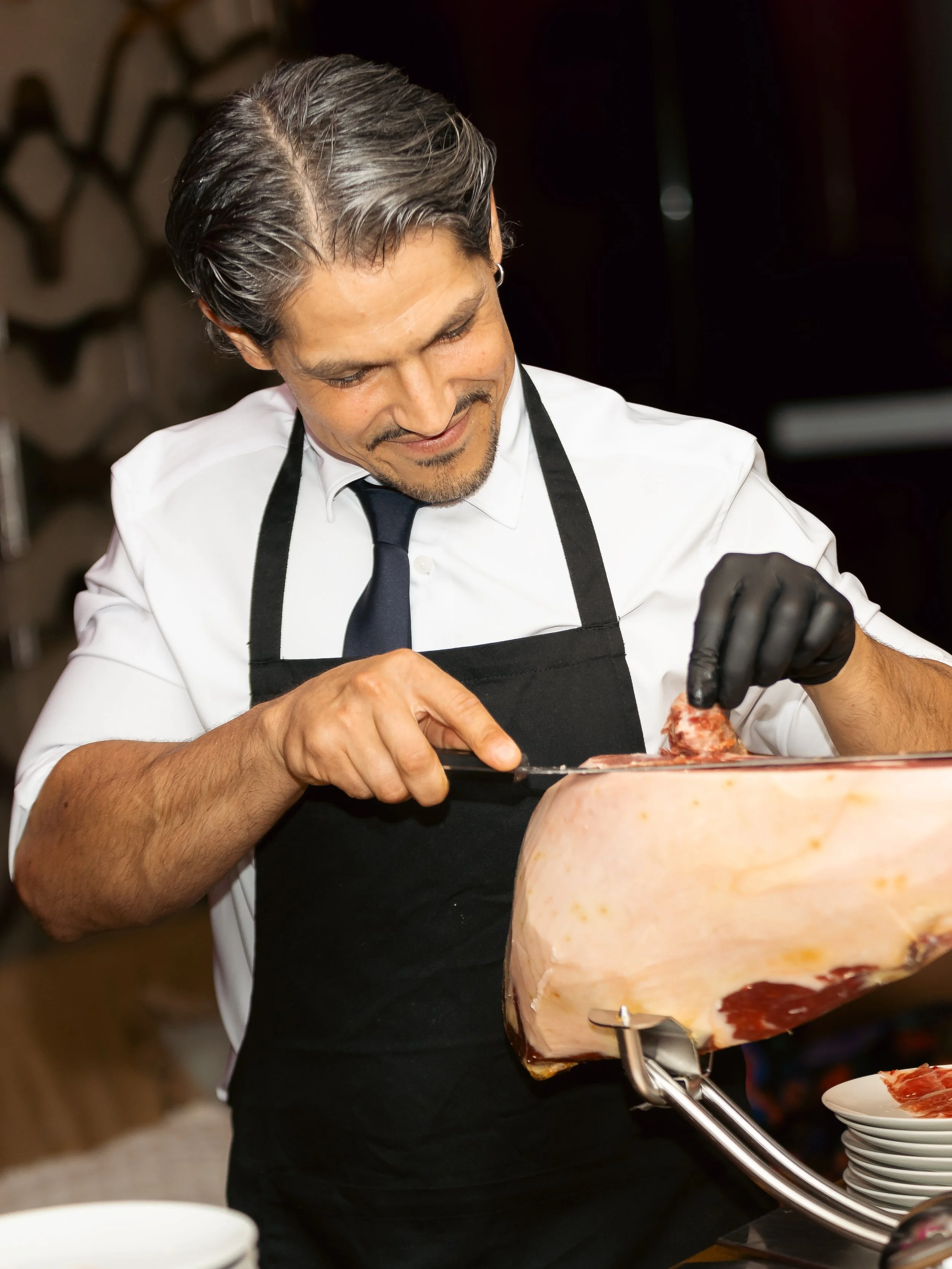A man with gray hair and a beard, dressed in a white shirt, black apron, and black gloves, is slicing cured meat, such as prosciutto, in a professional kitchen or deli.