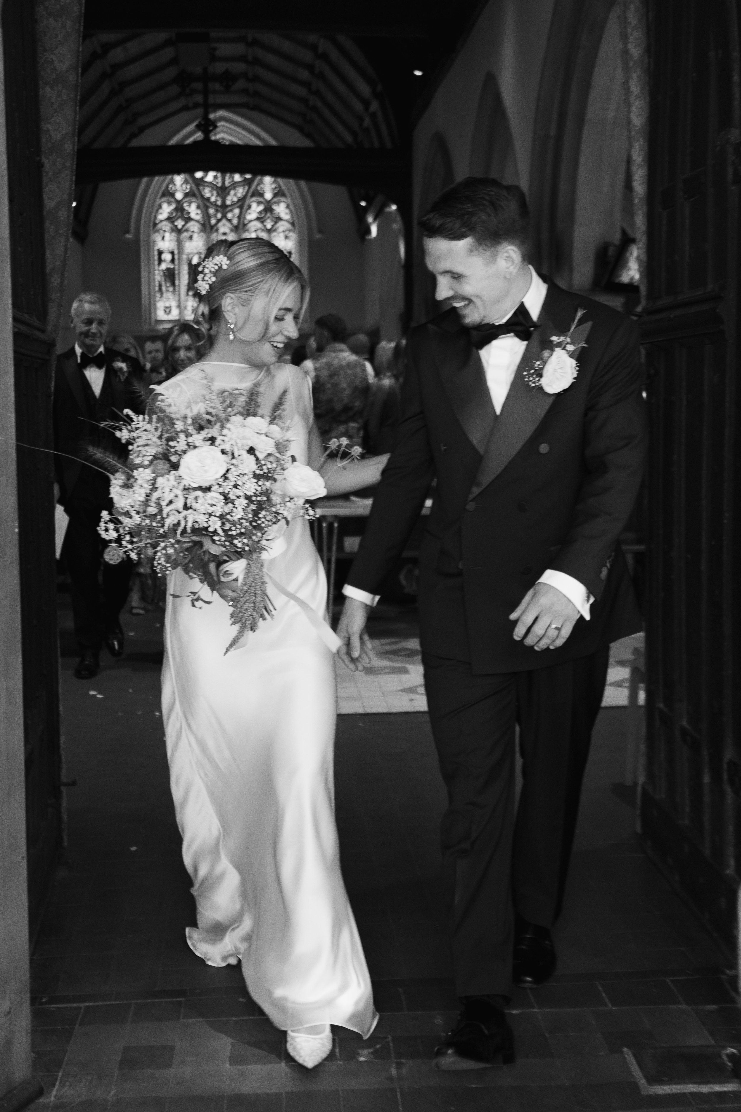 A bride and groom smiling and holding hands as they walk into a church for their wedding ceremony. The bride is wearing a white wedding dress and holding a bouquet of flowers, while the groom is in a black tuxedo with a bowtie.