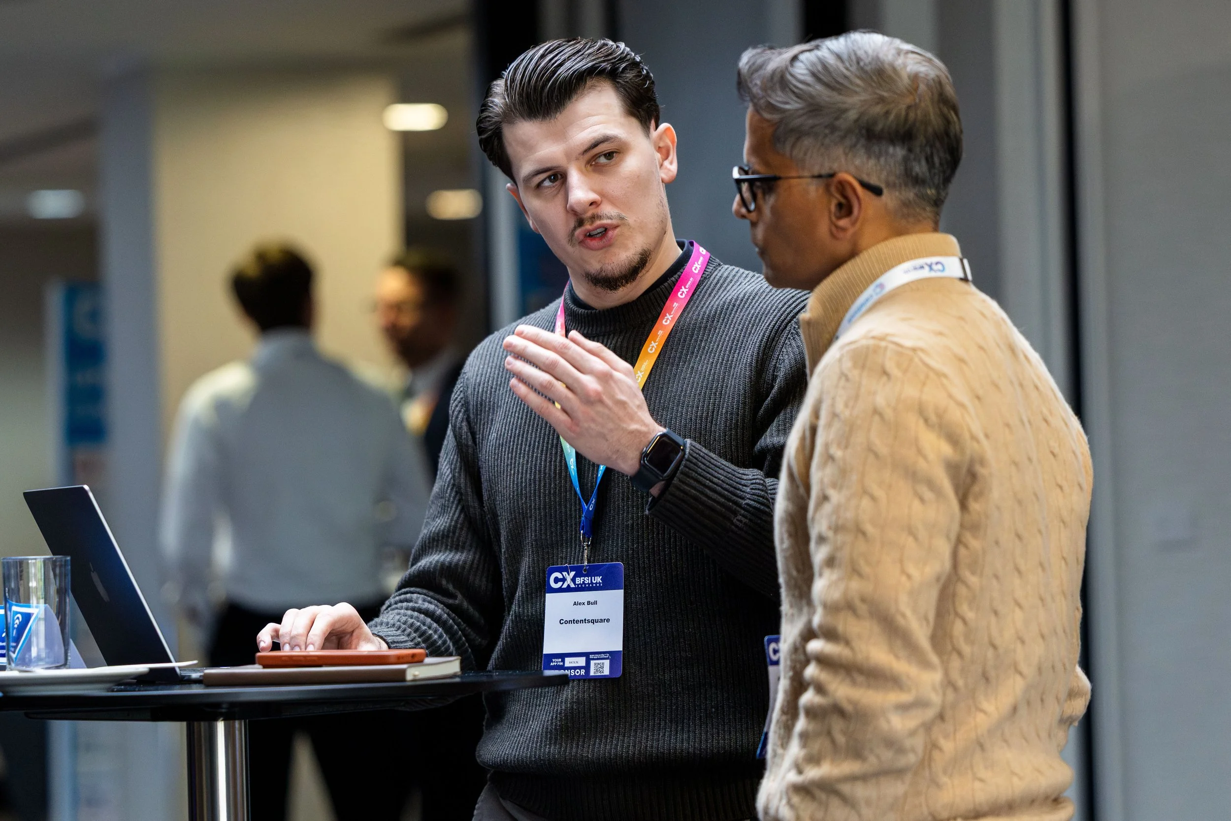 Two men engaged in conversation at a professional event, with one wearing a name tag labeled 'Alex Bull' and a lanyard, standing by a table with a laptop and drinks.