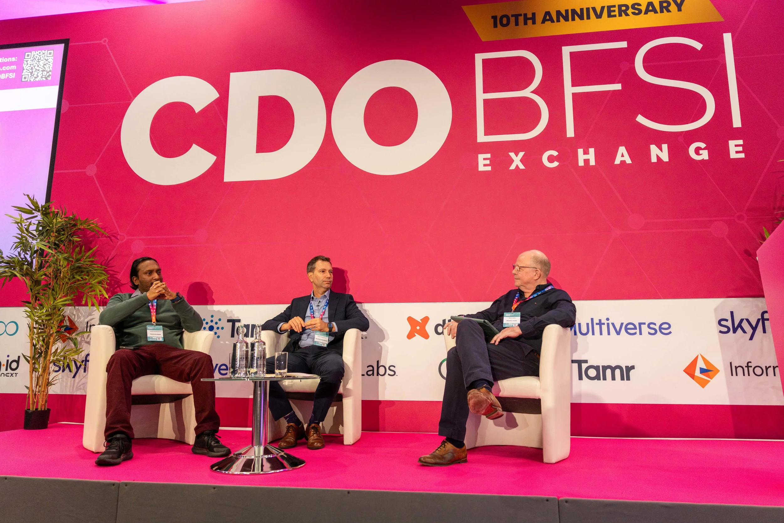 Three men seated on a stage participating in a panel discussion at a conference. Behind them is a large pink banner with the text '10th Anniversary CBDOES Exchange' and various sponsor logos. There are bottles of water on a small table in front of th