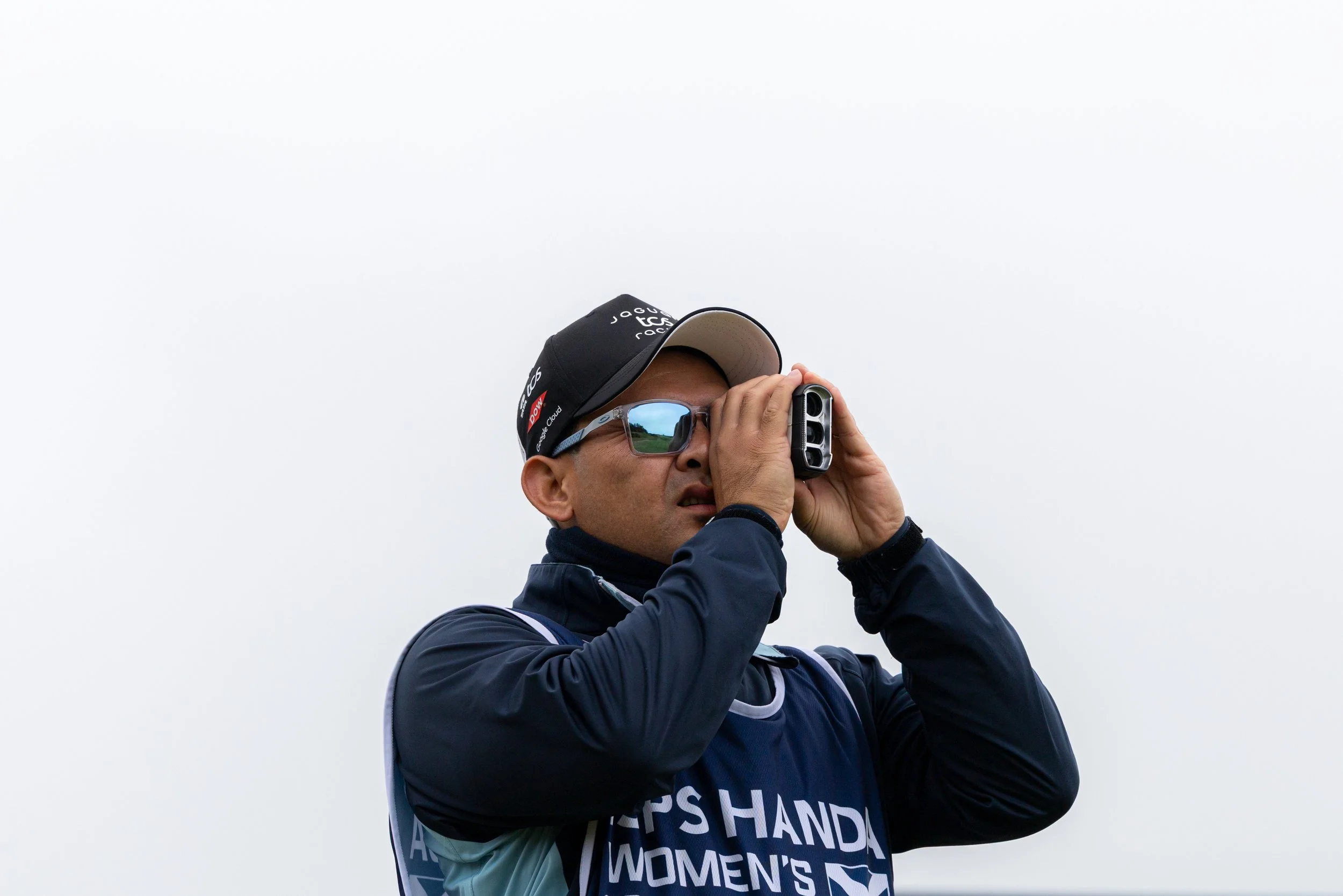 A person wearing sunglasses and a baseball cap, looking through binoculars or a camera, against a gray sky background.