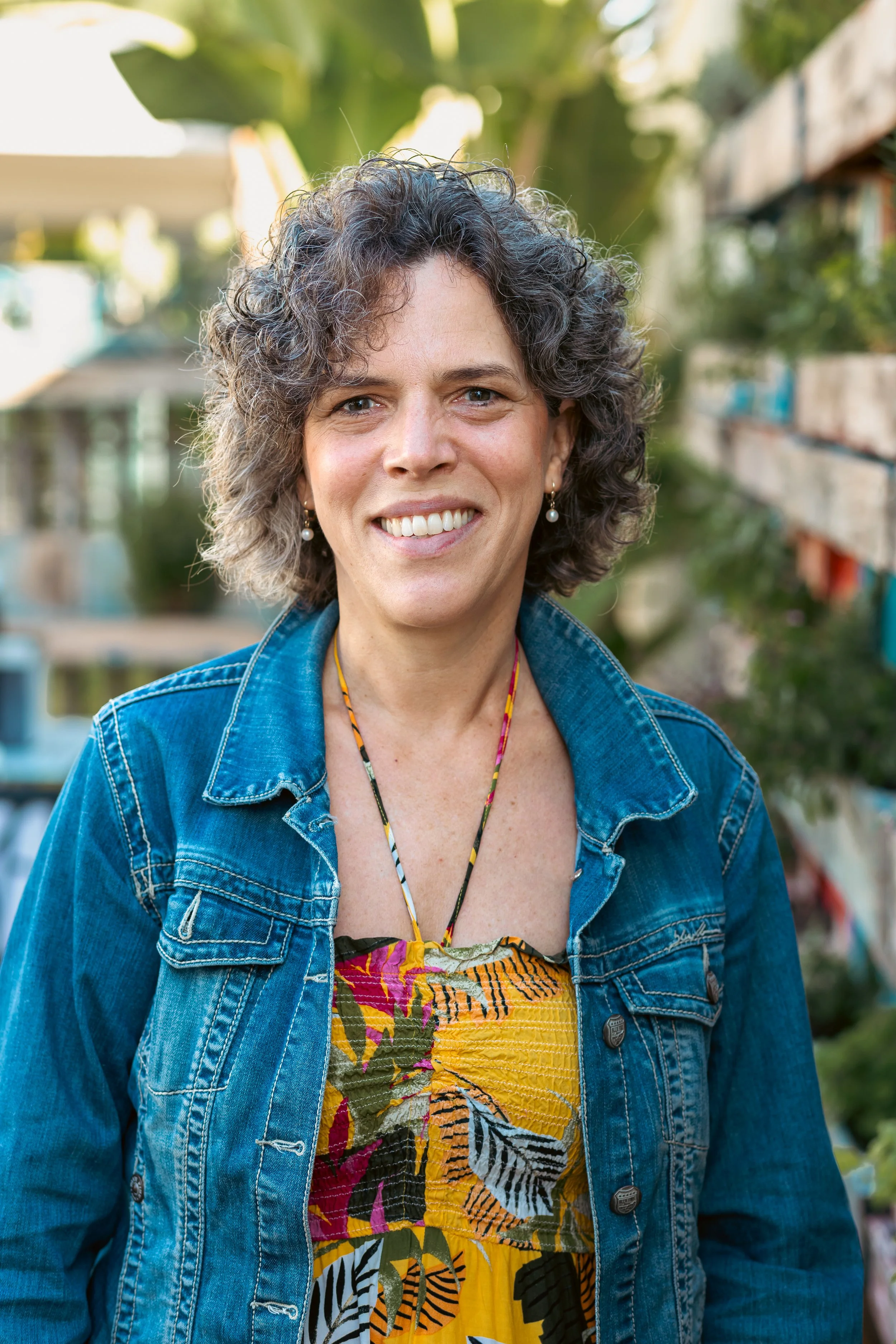 A woman with curly gray hair, wearing pearl earrings, a colorful patterned dress, and a denim jacket, smiling outdoors in front of a background of greenery and shelves.