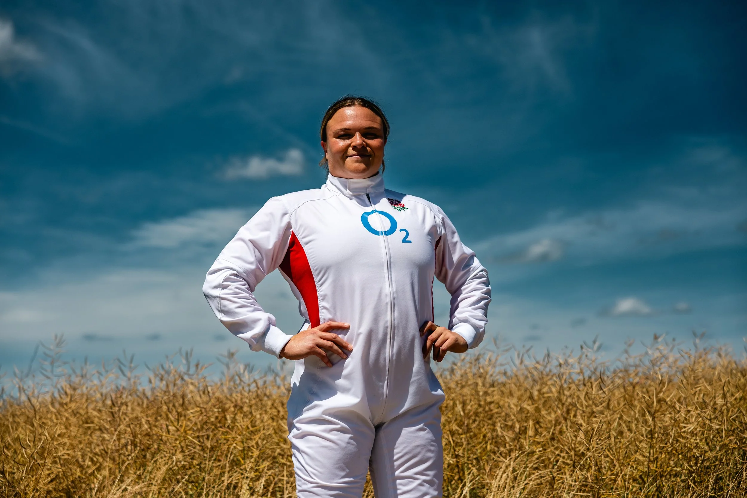 A woman in a white jacket with the London 2012 Olympics logo stands outdoors in a field of tall, dry grass under a partly cloudy sky.