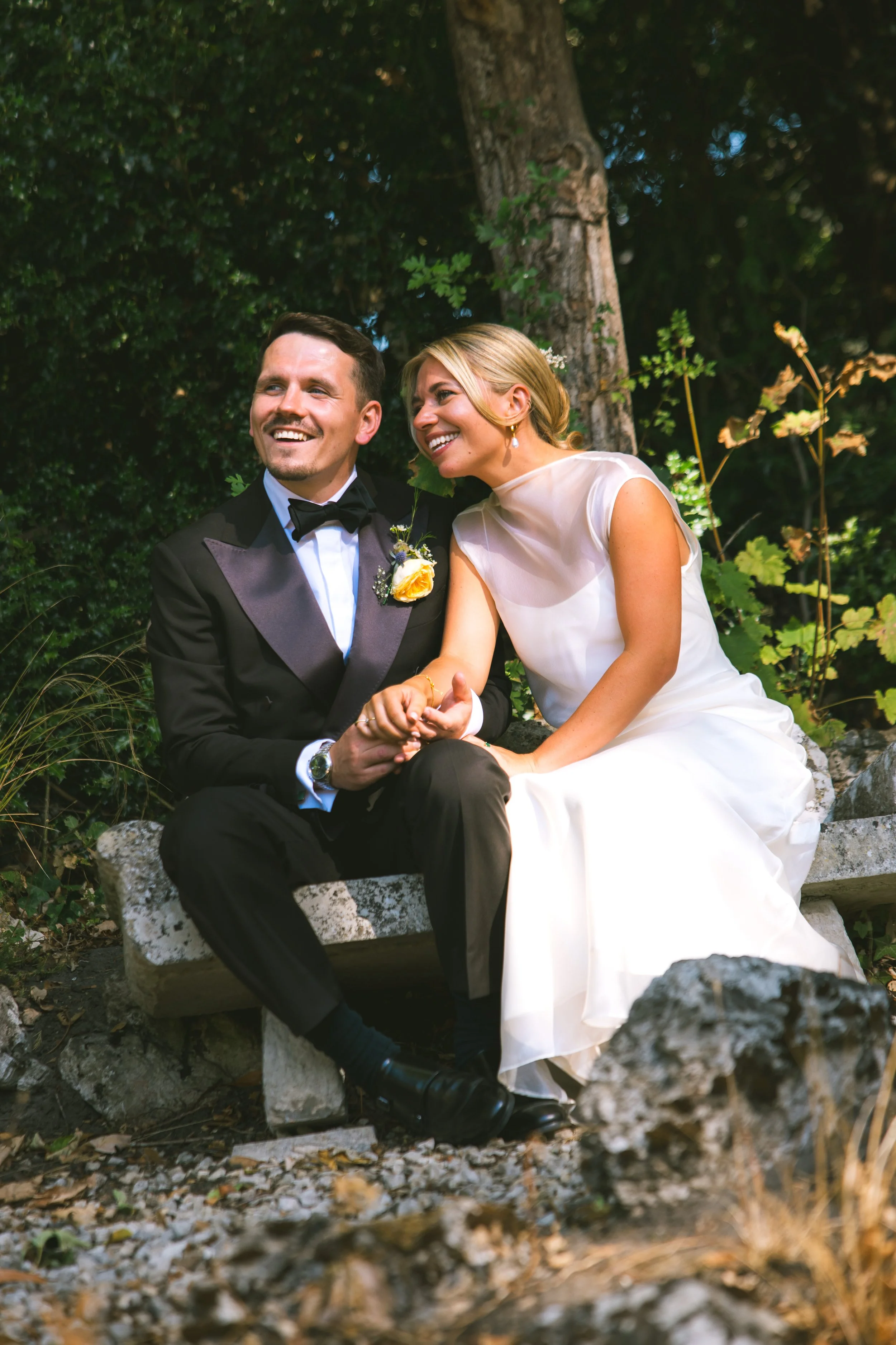 A bride and groom sitting on a stone bench outdoors, smiling and holding hands, surrounded by greenery and trees.