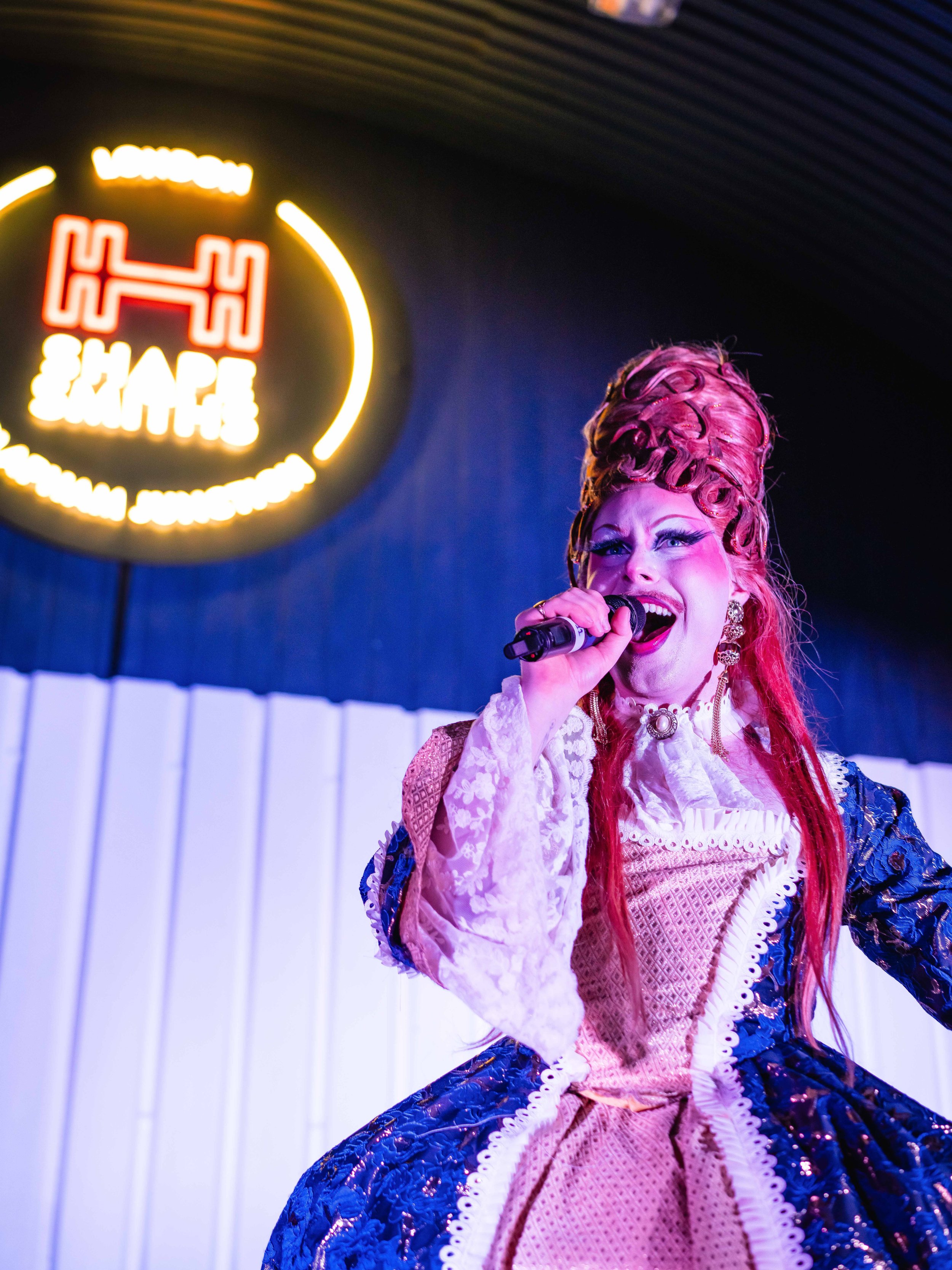 A woman dressed in colorful, elaborate costume with a tall, pink wig, singing into a microphone, with a neon sign in the background.