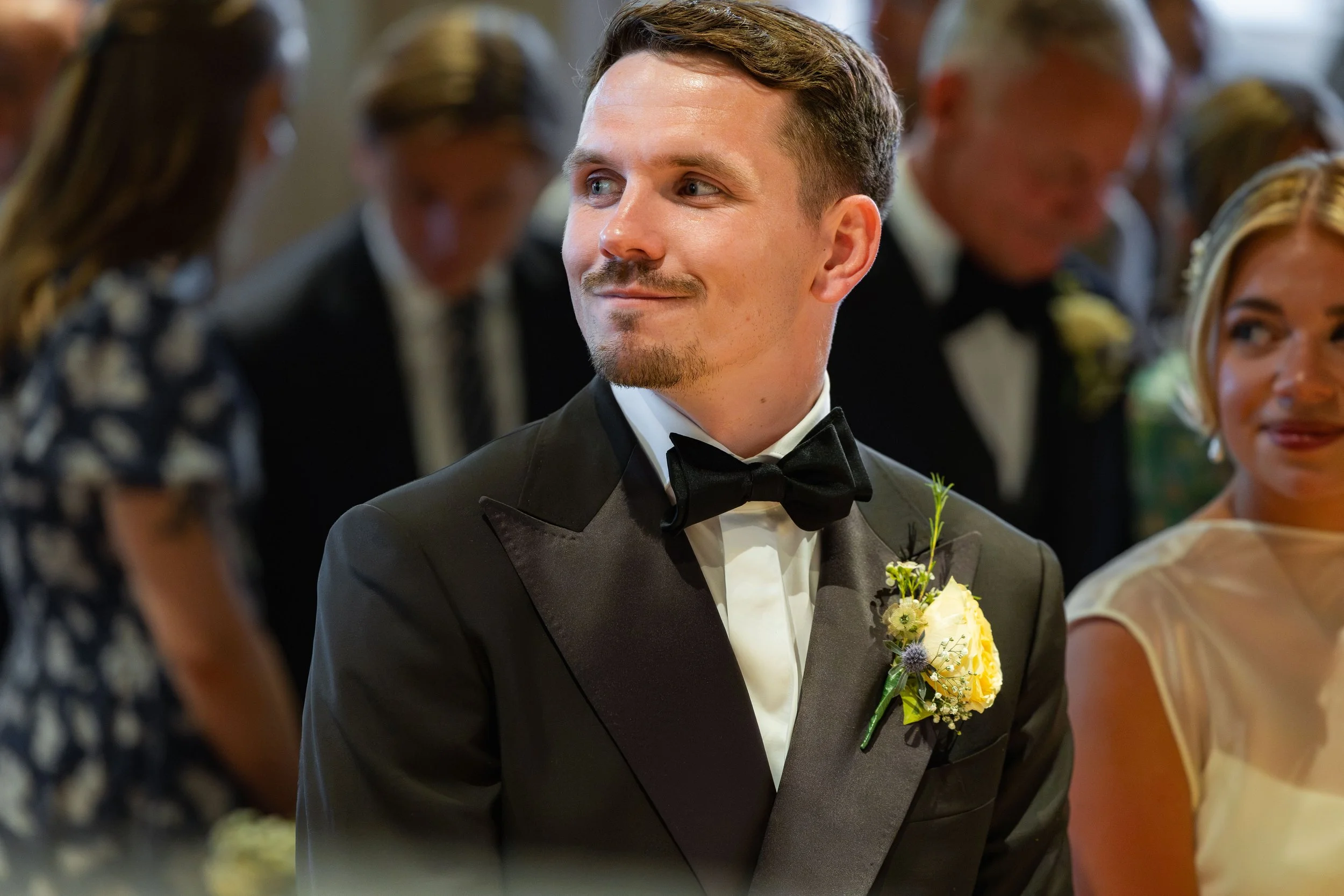 A man dressed in a black tuxedo with a bowtie, wearing a yellow boutonnière, is smiling and looking to his left during a wedding reception. He is surrounded by blurred people, some women in dresses and some men in tuxedos, in a festive indoor setting