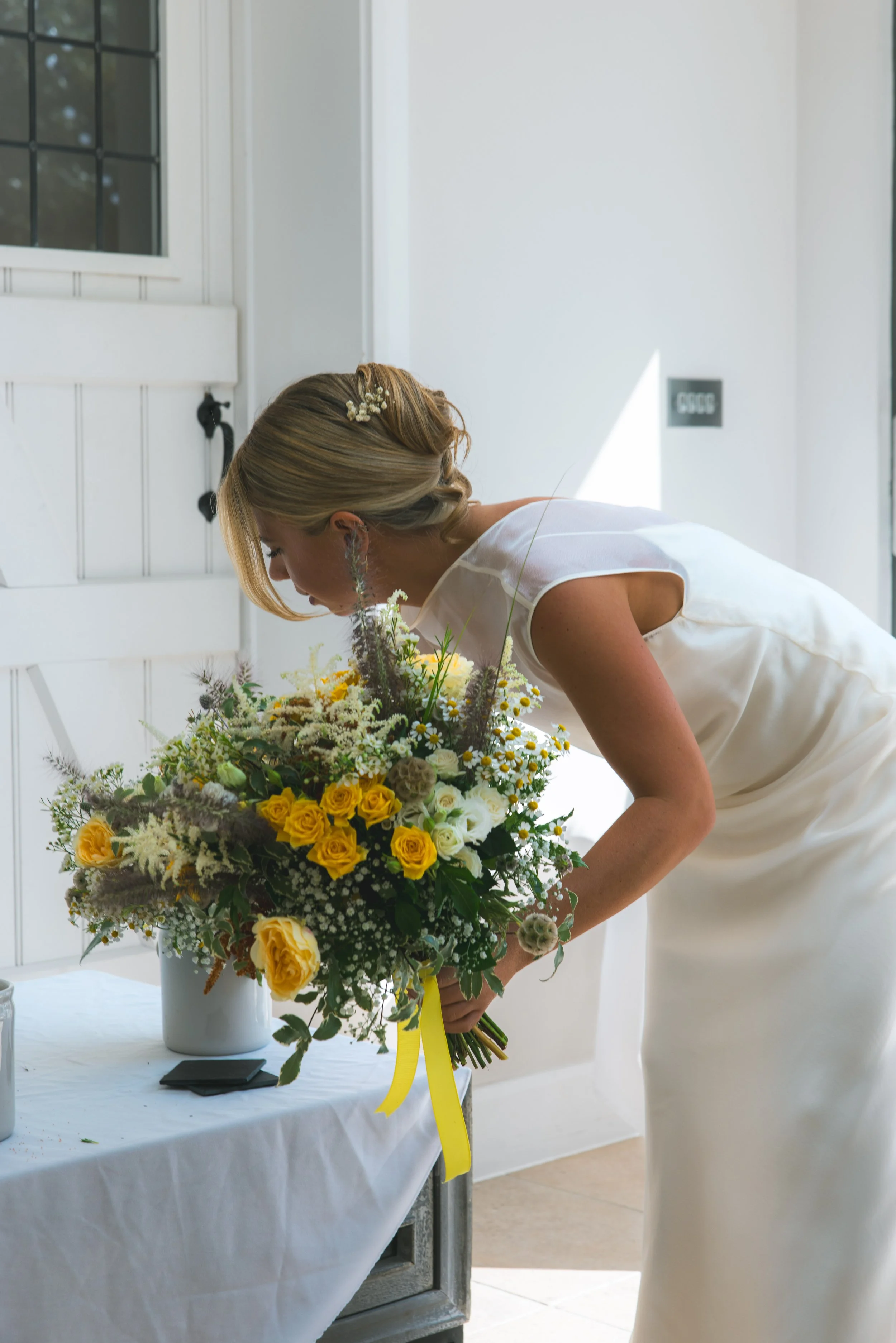 Woman in wedding dress placing a bouquet of yellow and white flowers on a table decorated with a white tablecloth during a wedding ceremony.