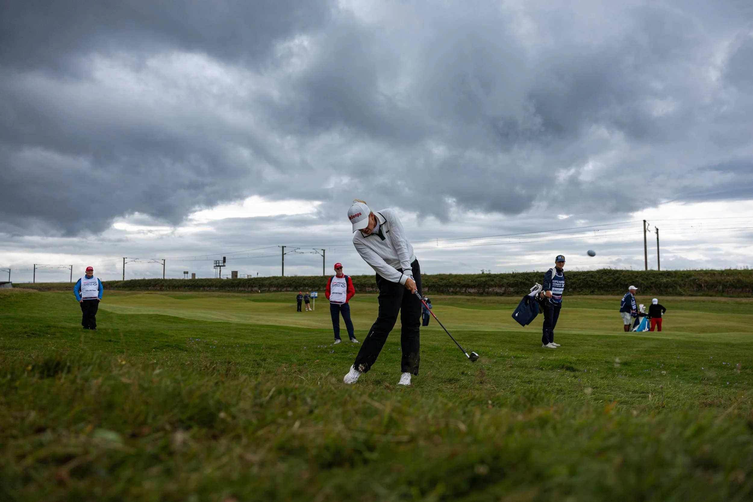 A group of people on a golf course under a cloudy sky. One person in foreground swings a golf club, others stand nearby with bags and equipment.