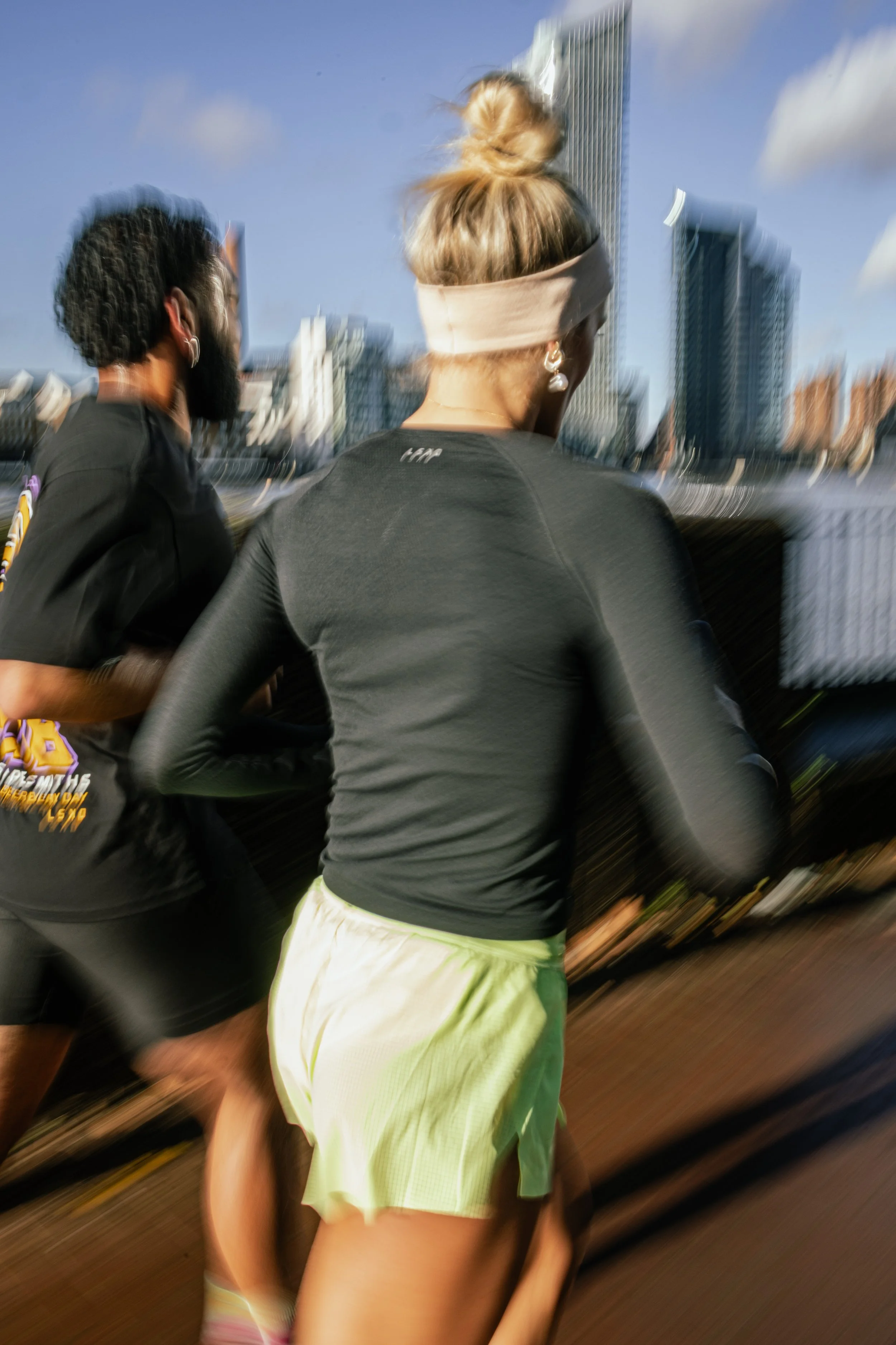 A woman running outdoors in athletic clothing with a cityscape background, wearing a headband, earrings, and shorts.