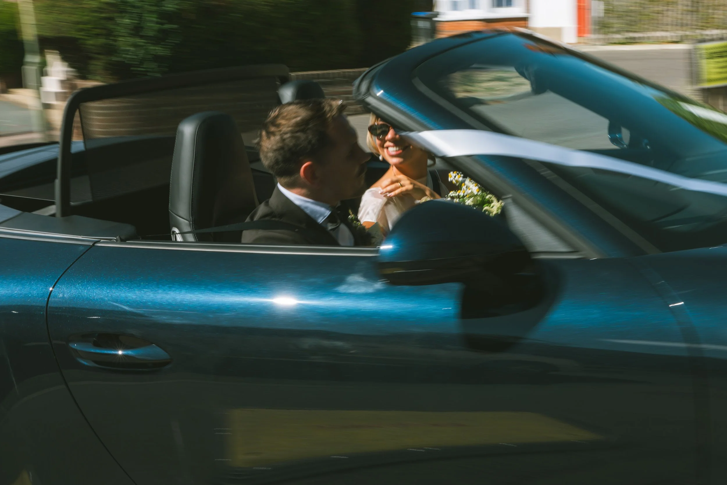 A couple in a convertible car, with the woman smiling and wearing sunglasses, driving during daytime with greenery in the background.