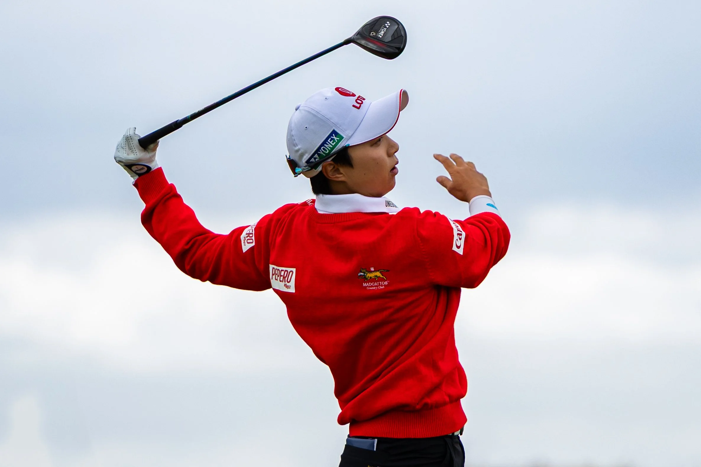 A golfer in a red jacket and white cap swinging a golf club against a cloudy sky.