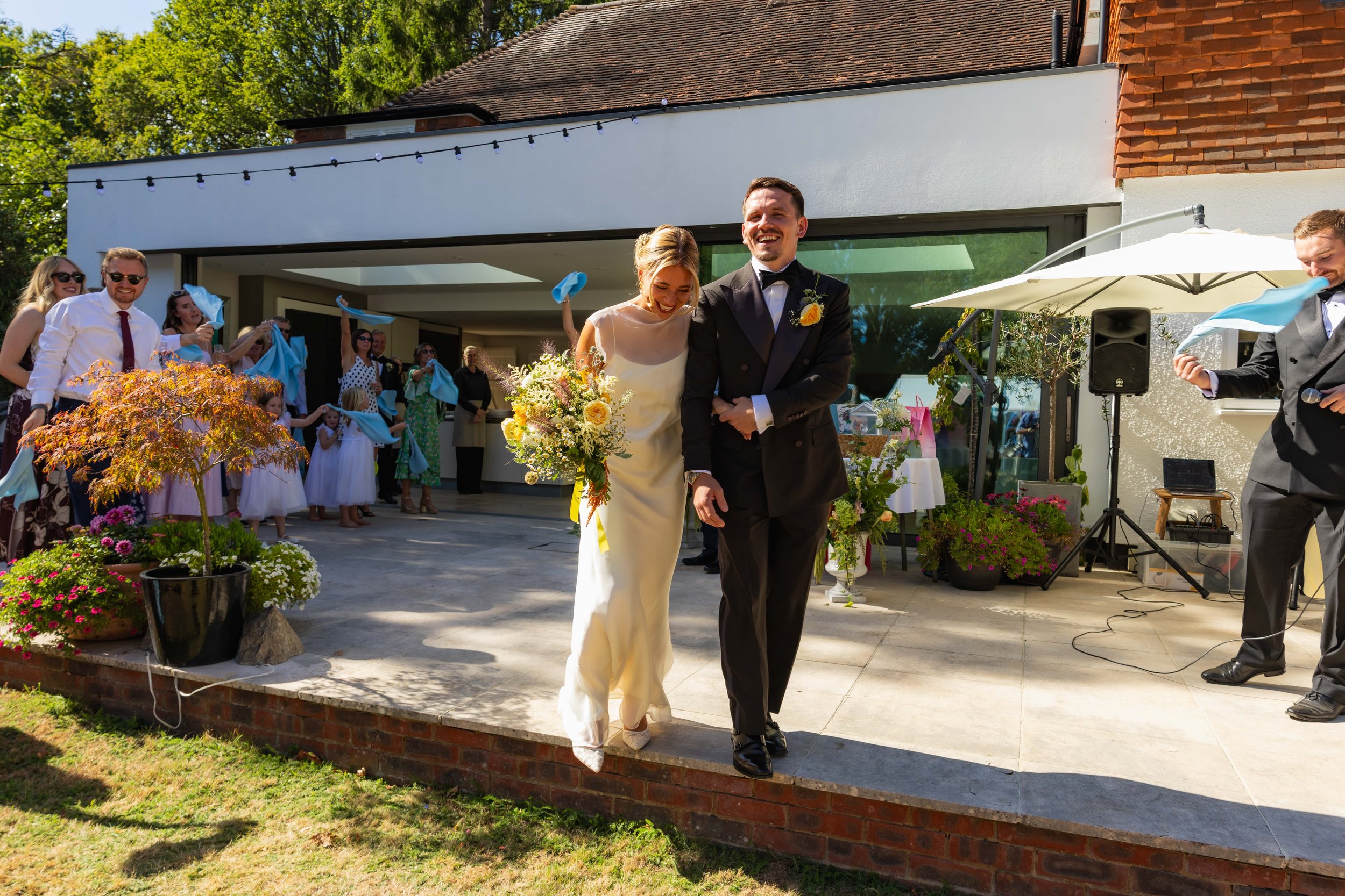 A wedding celebration outdoors with a bride and groom walking together. The bride is holding a bouquet and the groom is dressed in a tuxedo. Guests are in the background, some waving blue cloths, on a patio decorated with potted plants, flowers, and 