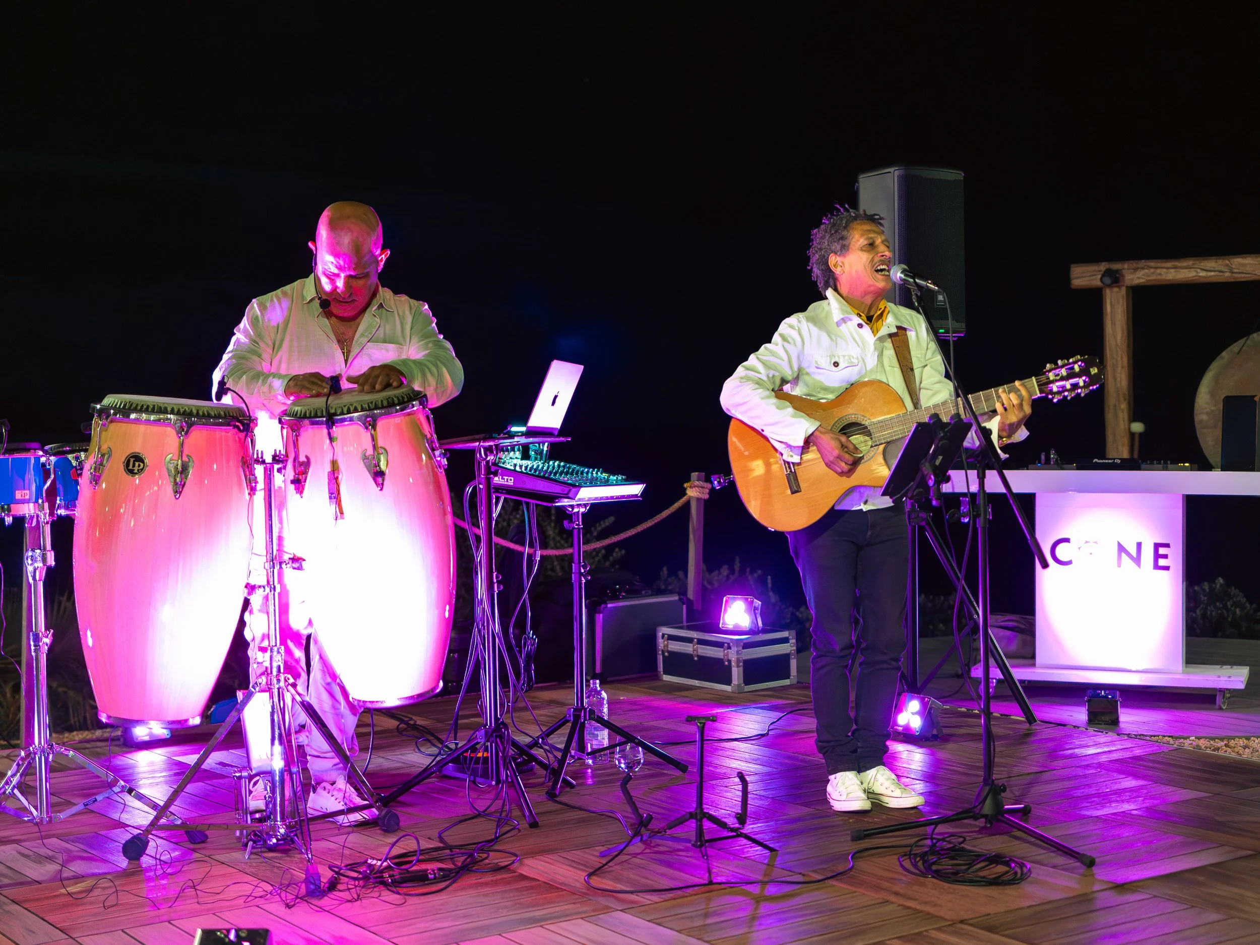 Two musicians performing outdoors at night, one playing conga drums and the other singing and playing an acoustic guitar, with colorful stage lighting.