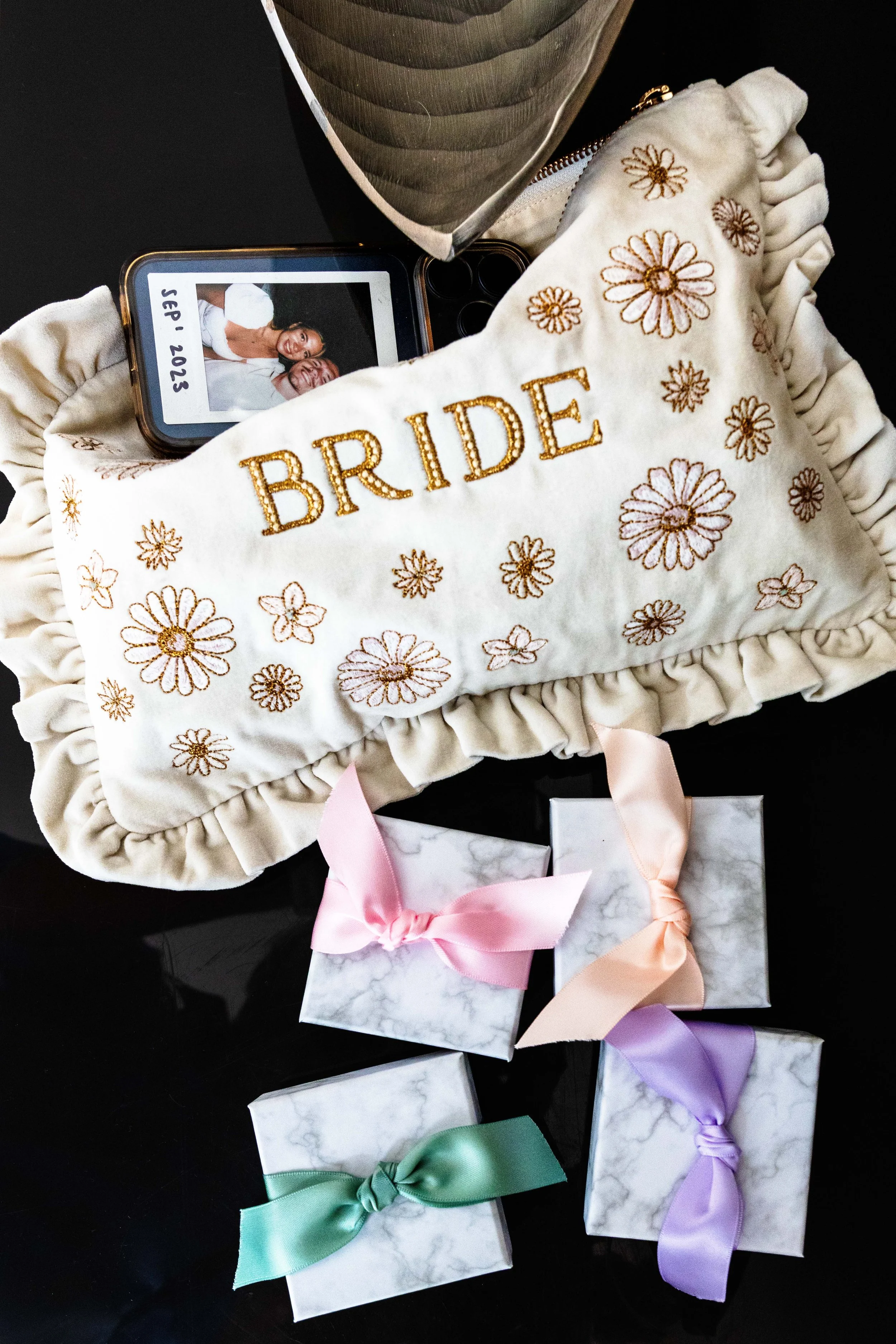 A collection of small gift boxes wrapped in white marbled paper with pastel-colored ribbons (green, pink, purple) and bows, arranged on a black surface. There is also an embroidered bridal sash with the word "BRIDE" and daisy flower patterns, a photo