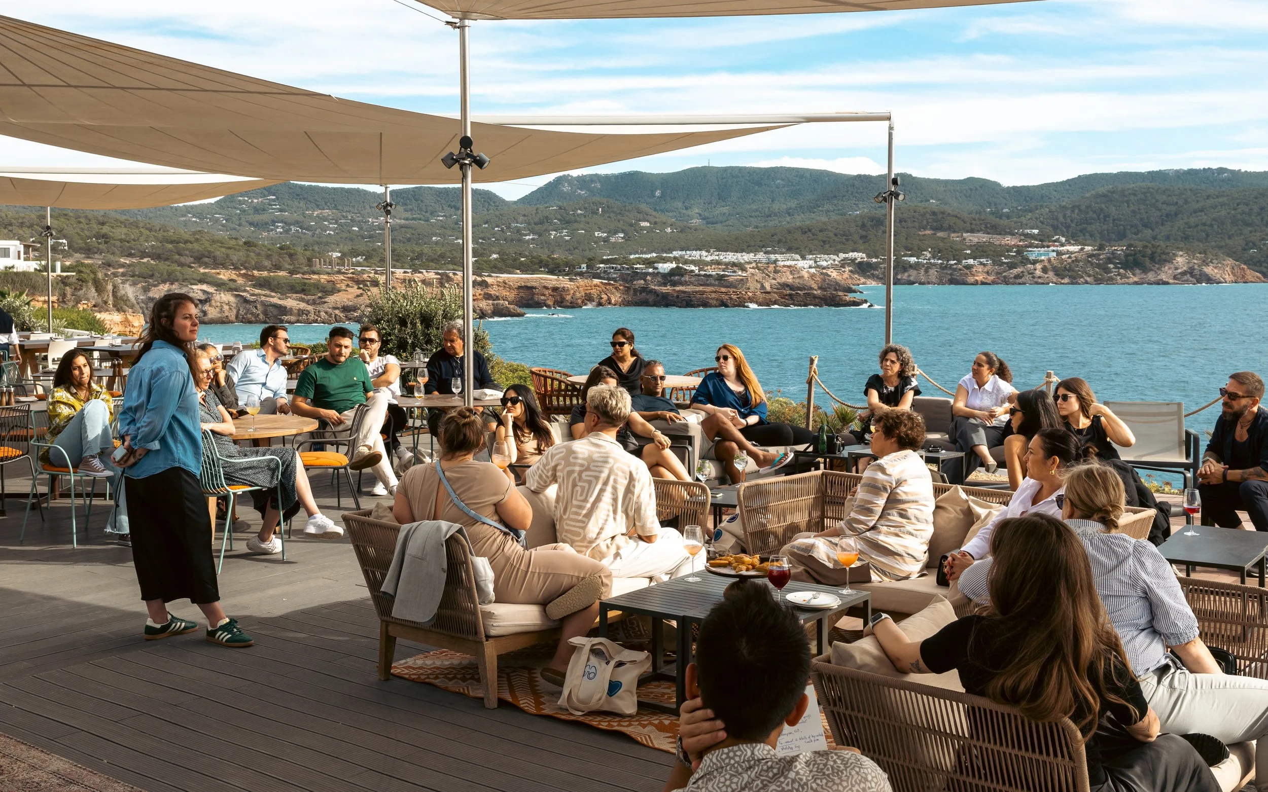 A group of people gathered on a patio overlooking the ocean with rocky cliffs and ocean in the background, sitting at tables and listening to a woman speak.