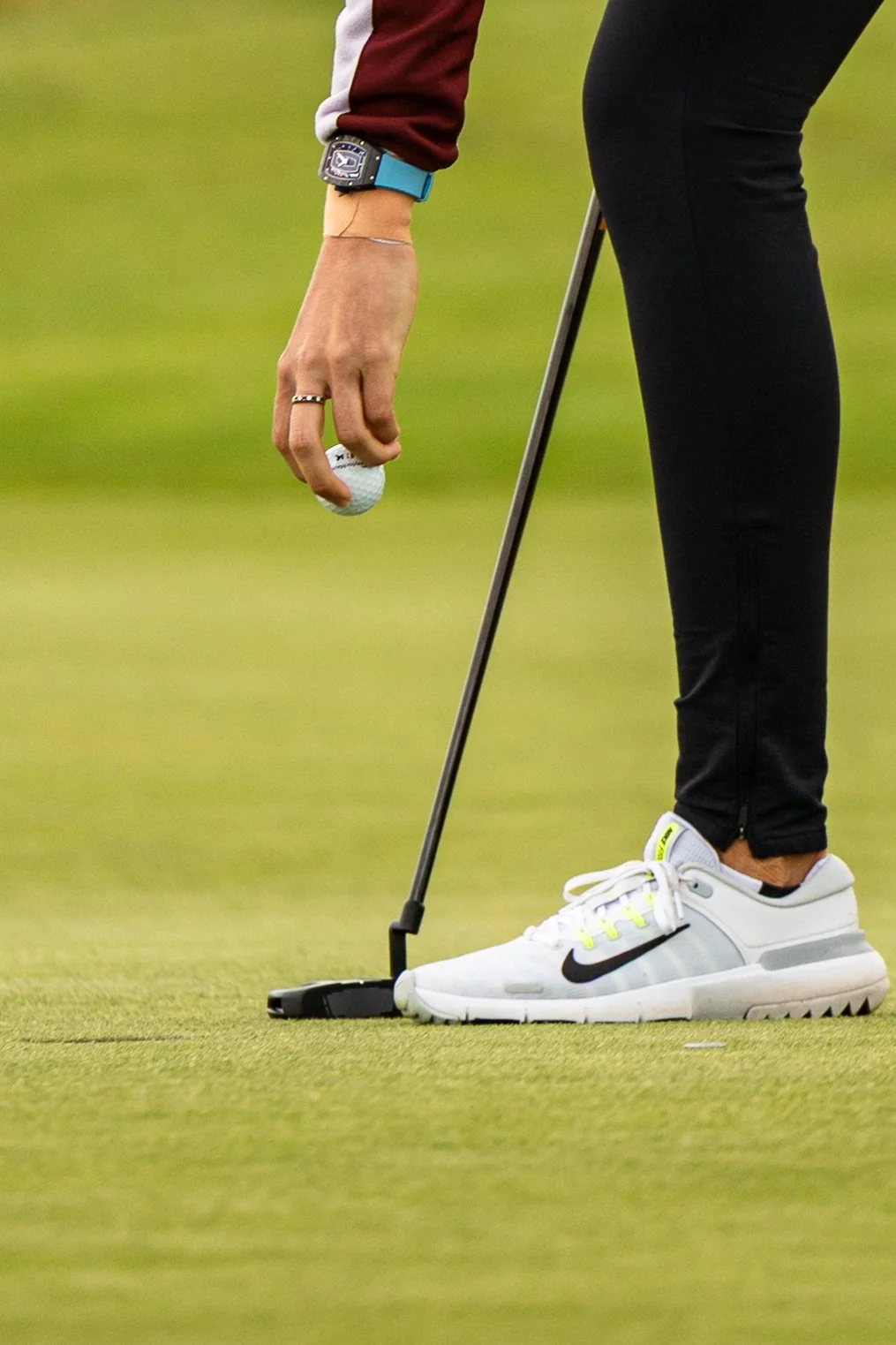 A person holding a golf ball on a putting green, wearing white Nike golf shoes, black pants, and a long sleeve shirt, with a watch on the left wrist and a ring on the left hand, using a golf club.
