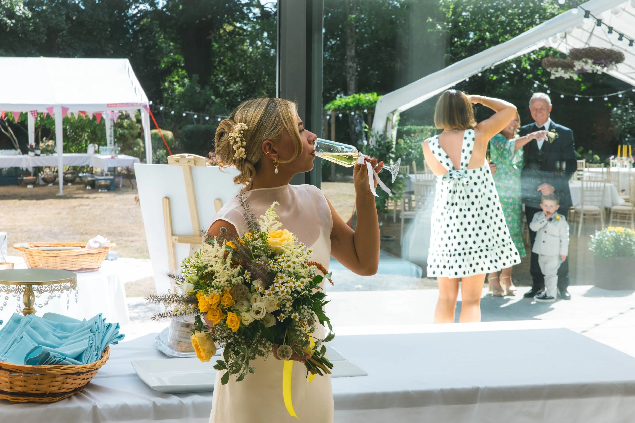 A woman in a white dress with floral details holding a bouquet of yellow and white flowers, sipping a glass of champagne, with a wedding reception in the background. Guests are celebrating outside with trees, tents, and decorations.