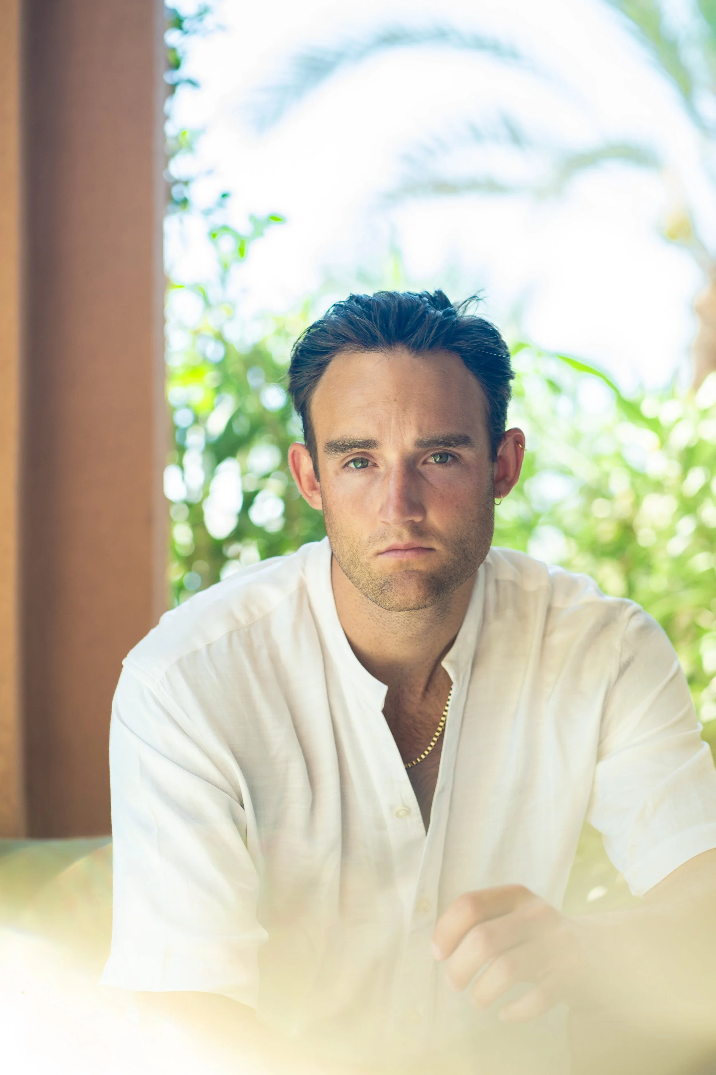 A man with dark hair styled back, wearing a white shirt with the top buttons undone, sitting indoors with a blurred background of green foliage and natural light.