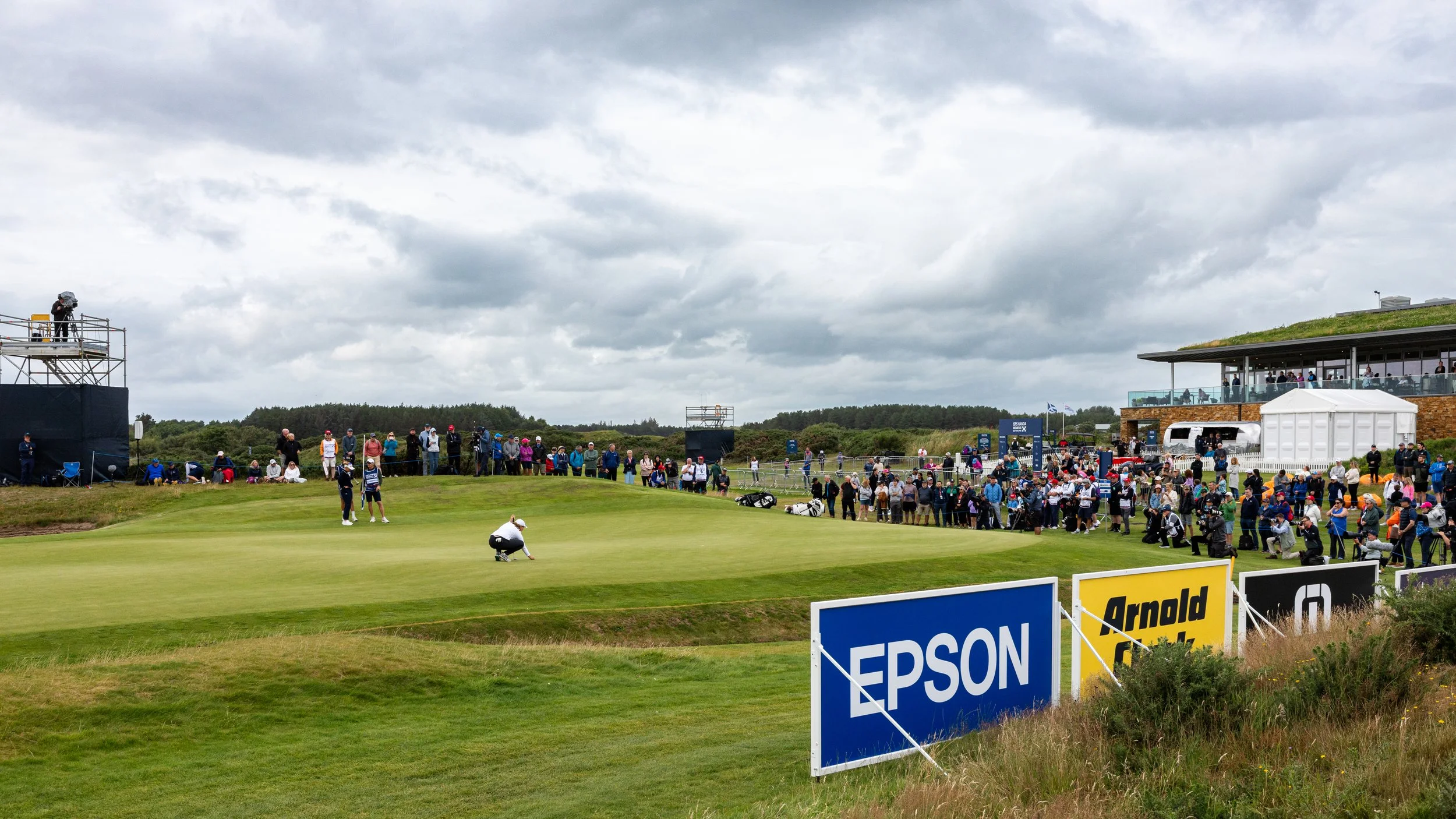 A golf course with a golfer preparing to putt on the green, surrounded by spectators and media, under a cloudy sky.
