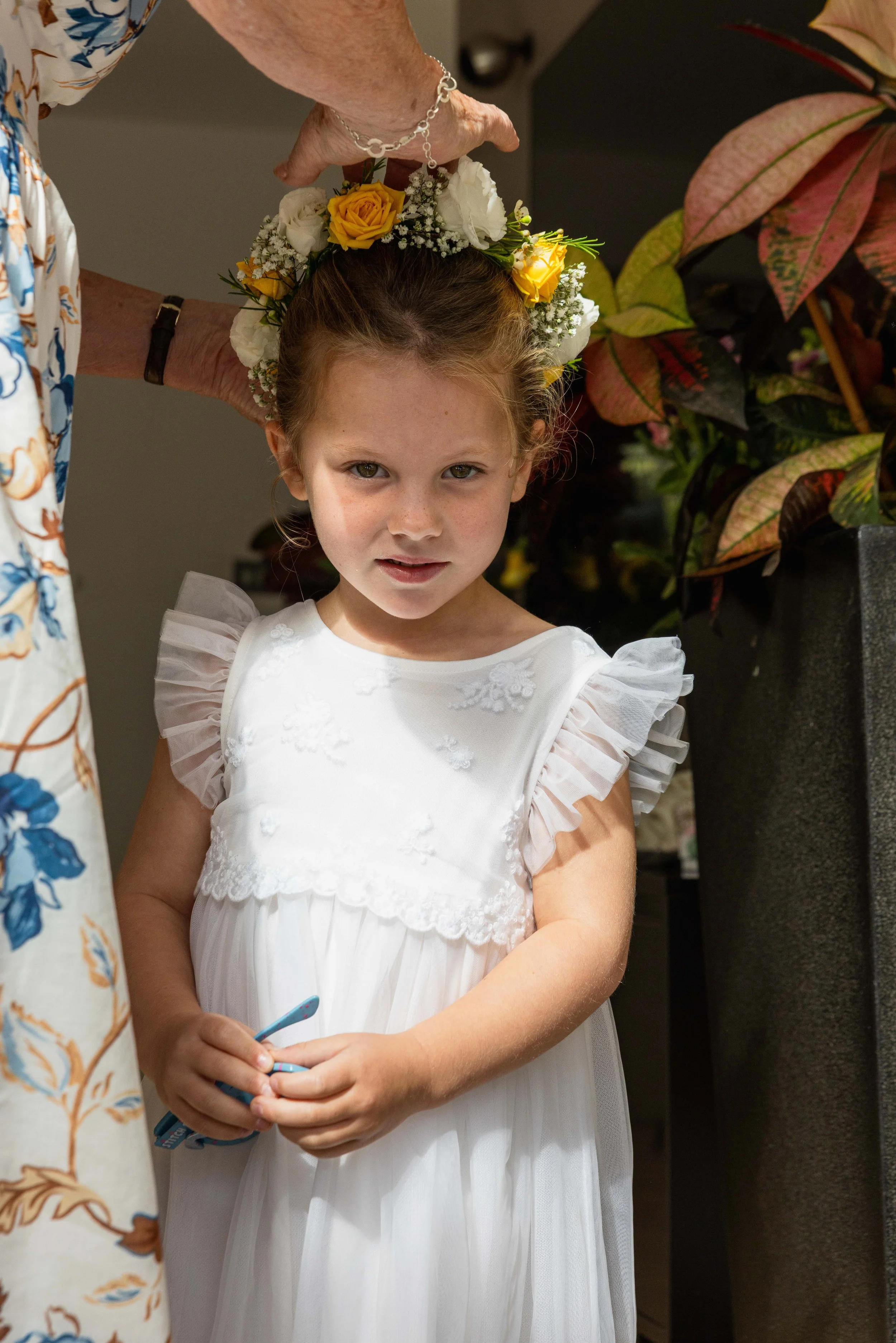 A young girl standing indoors, dressed in a white dress with ruffled sleeves, and wearing a floral headpiece made of yellow and white roses with small white filler flowers. An elderly person above her is adjusting the flower crown, with plants and a 