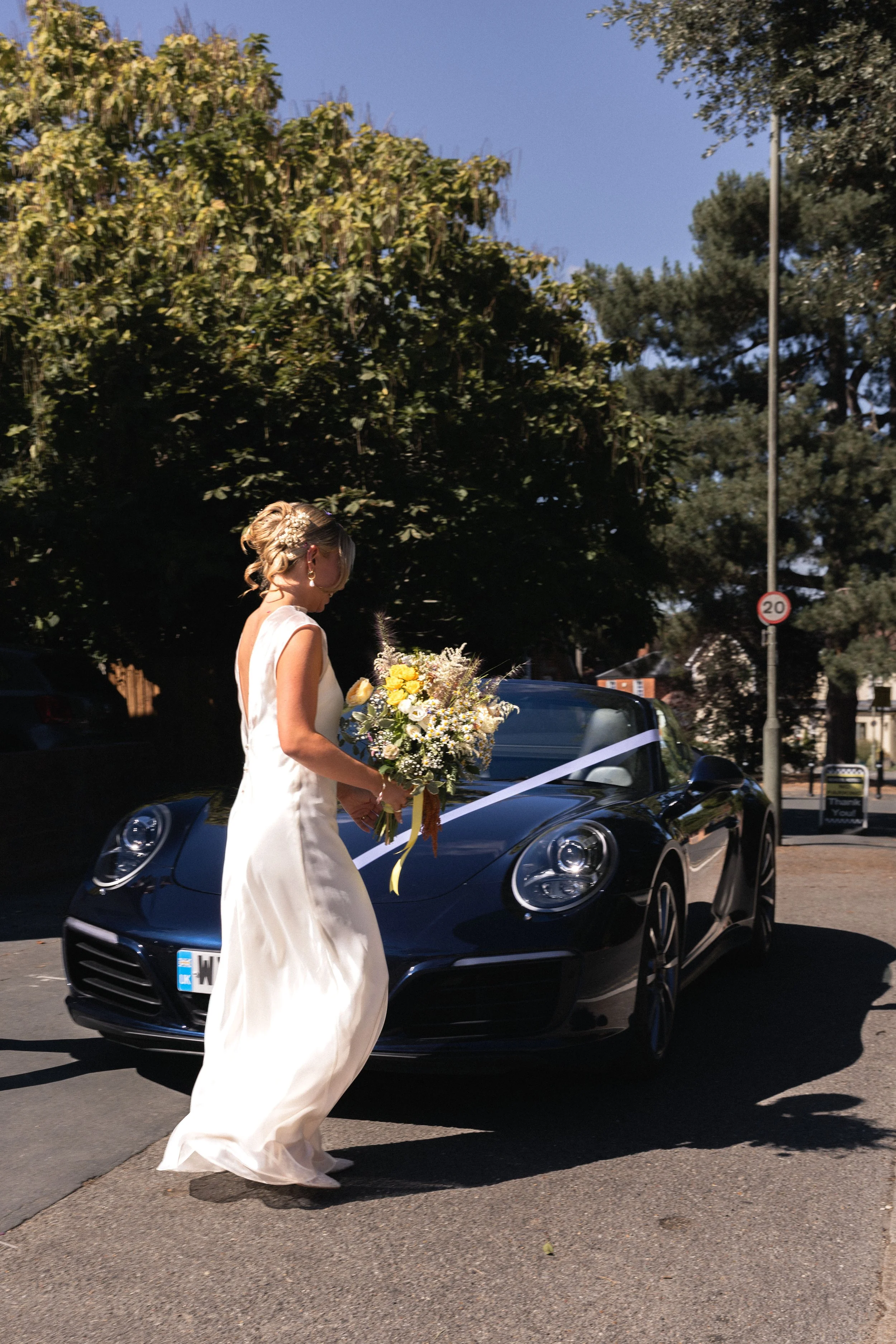 A woman in a white wedding dress holding a bouquet of flowers standing next to a dark blue luxury car with a white ribbon on it, outdoors on a sunny day with trees in the background.