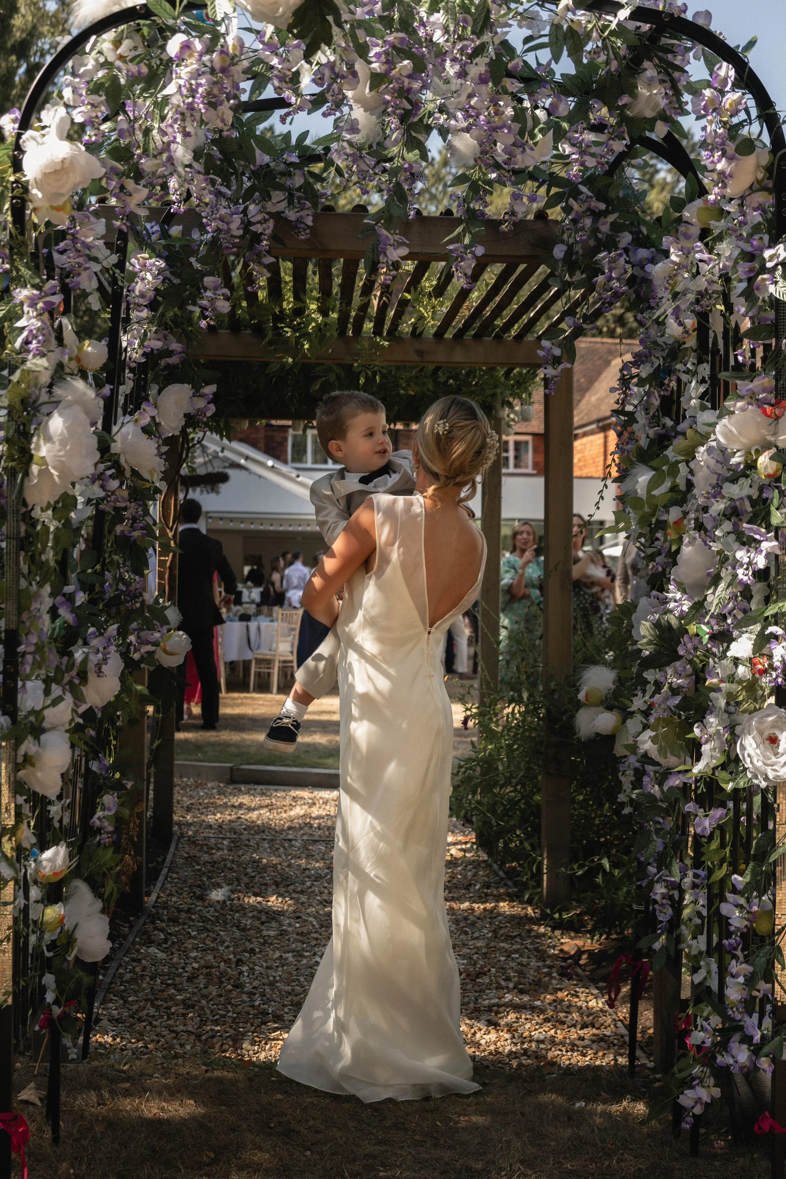A woman in a wedding dress holding a young boy in a tuxedo in an outdoor wedding setting. They are under a floral arch decorated with purple and white flowers, with guests in the background.