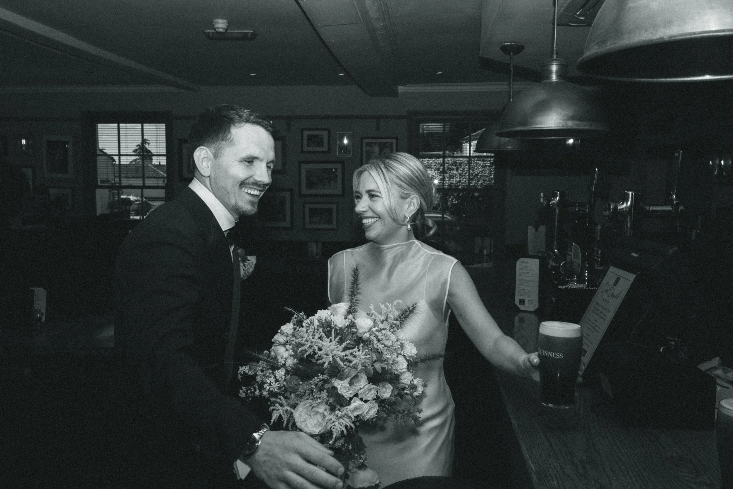 A black and white photo of a bride and groom smiling at each other, with the bride holding a bouquet of flowers. They appear to be celebrating, possibly at a wedding reception, with drinks on the table in front of them.