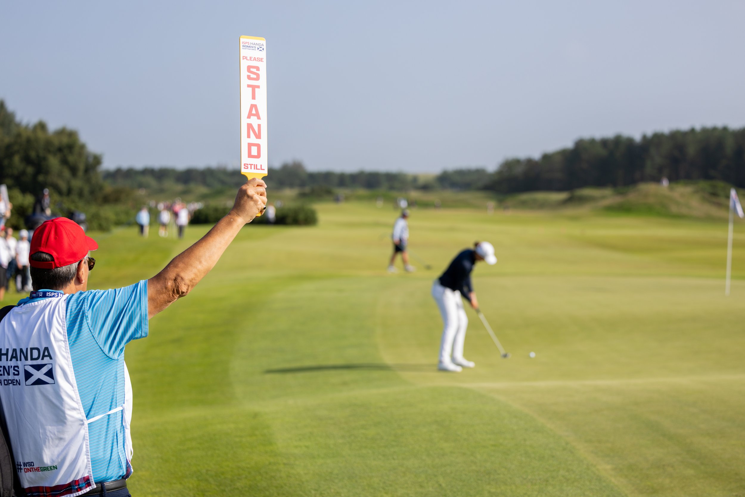 A person holding a sign that reads 'Please Stand Still' at a golf course during a golf tournament. The individual is wearing a white vest with blue and black accents, and a red cap. In the background, other golfers are preparing to take their shots o