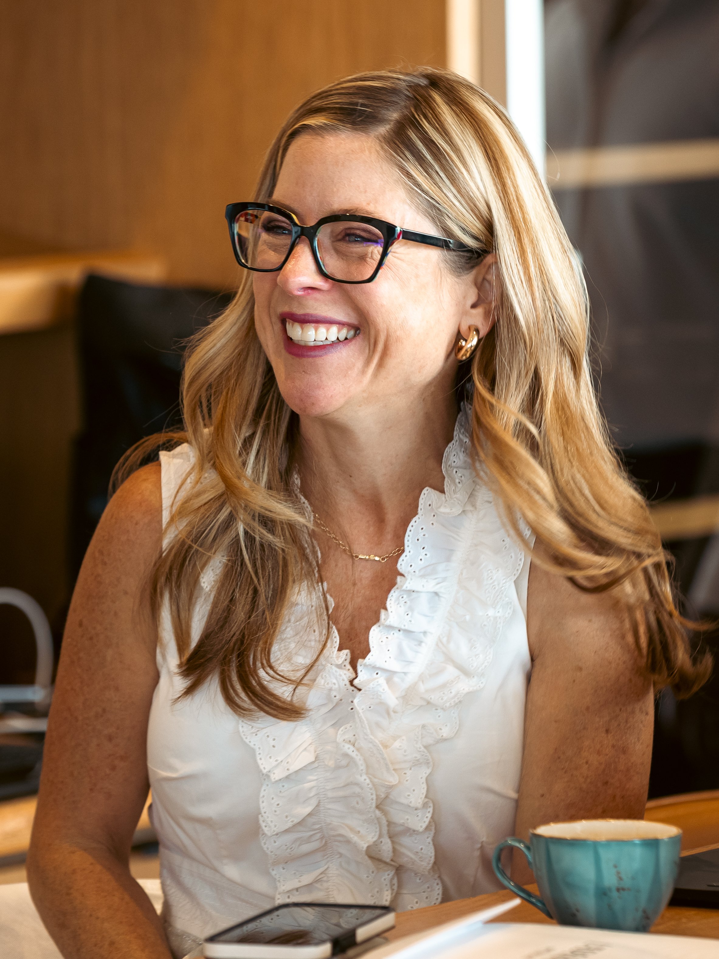 A smiling woman with long blonde hair, wearing black-rimmed glasses, gold hoop earrings, and a white sleeveless top with ruffle details, sitting at a table with a Teal mug, a smartphone, and a notebook in front of her, in a cozy indoor setting with w